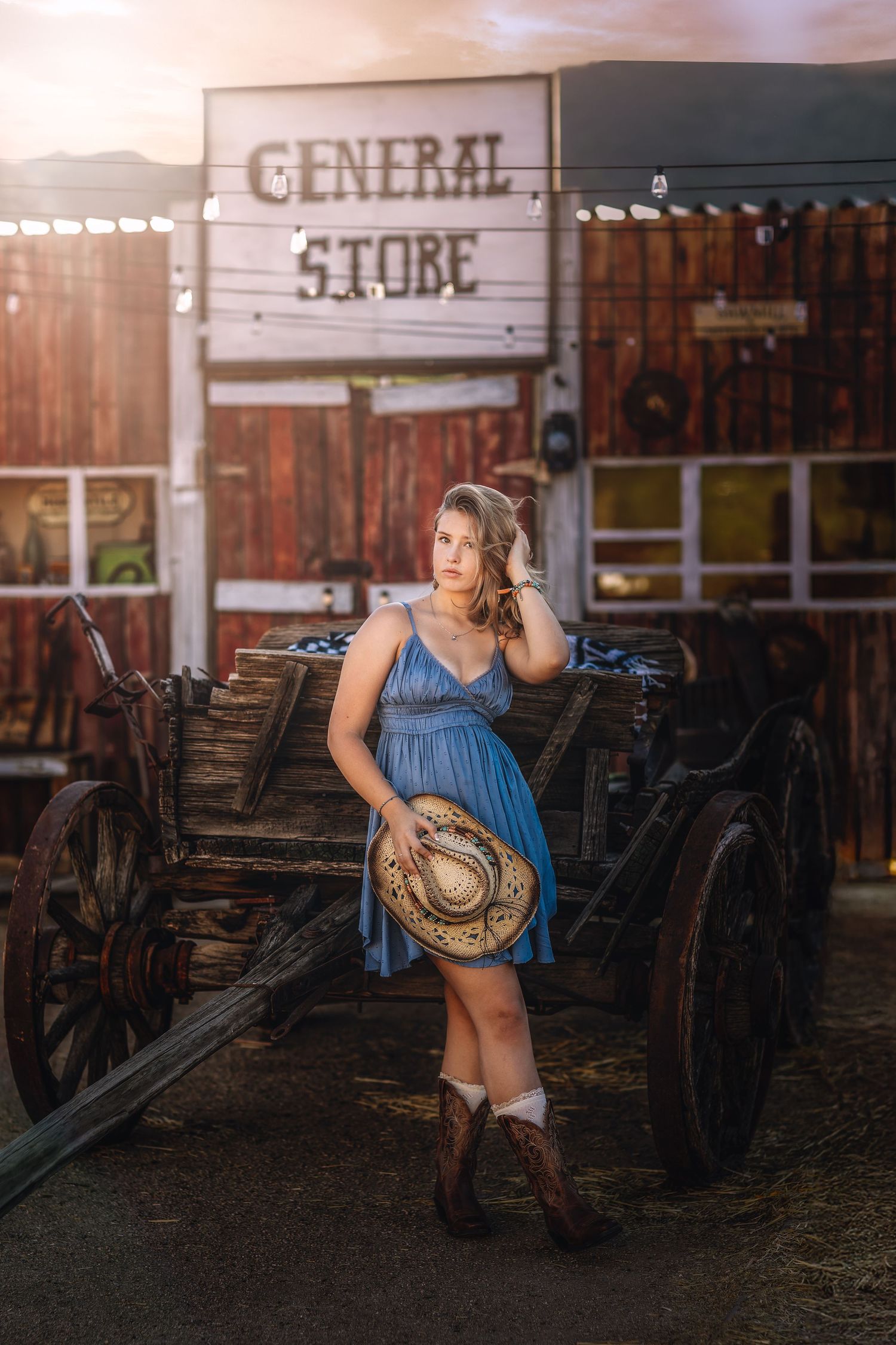 Young woman in blue dress with cowboy hat leaning on antique wagon by General Store at sunset.