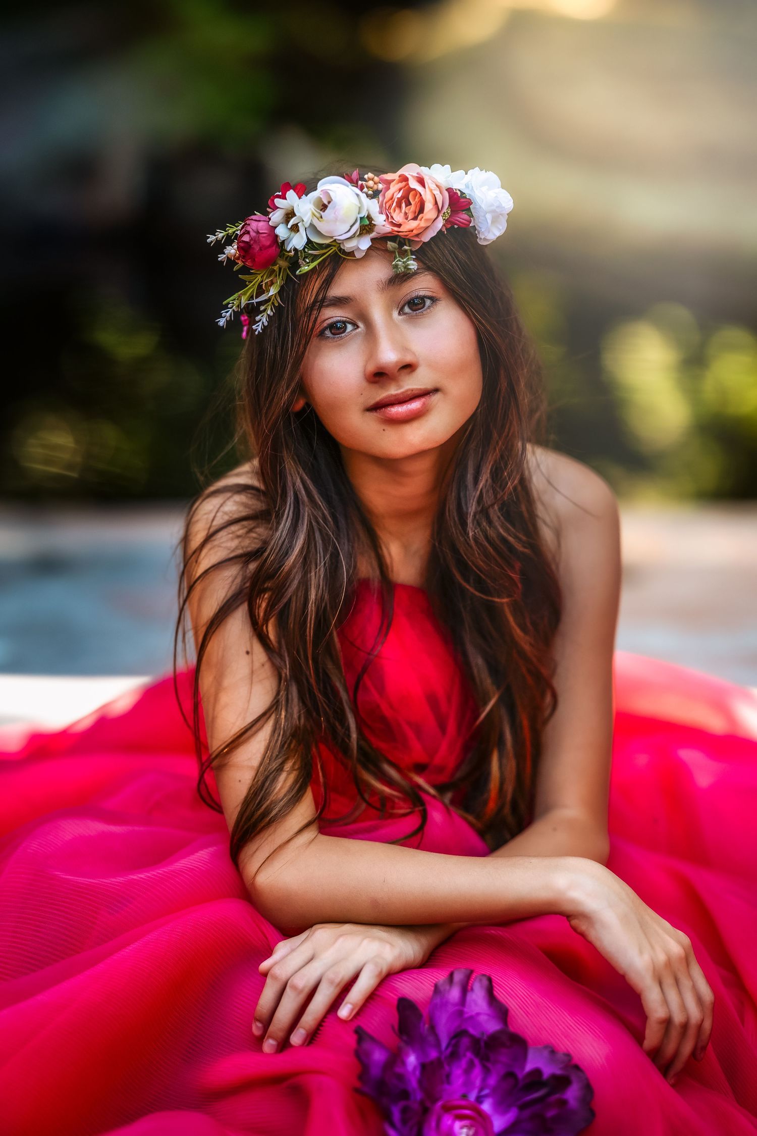 Teen girl in red dress wearing floral crown poses outdoors in garden.