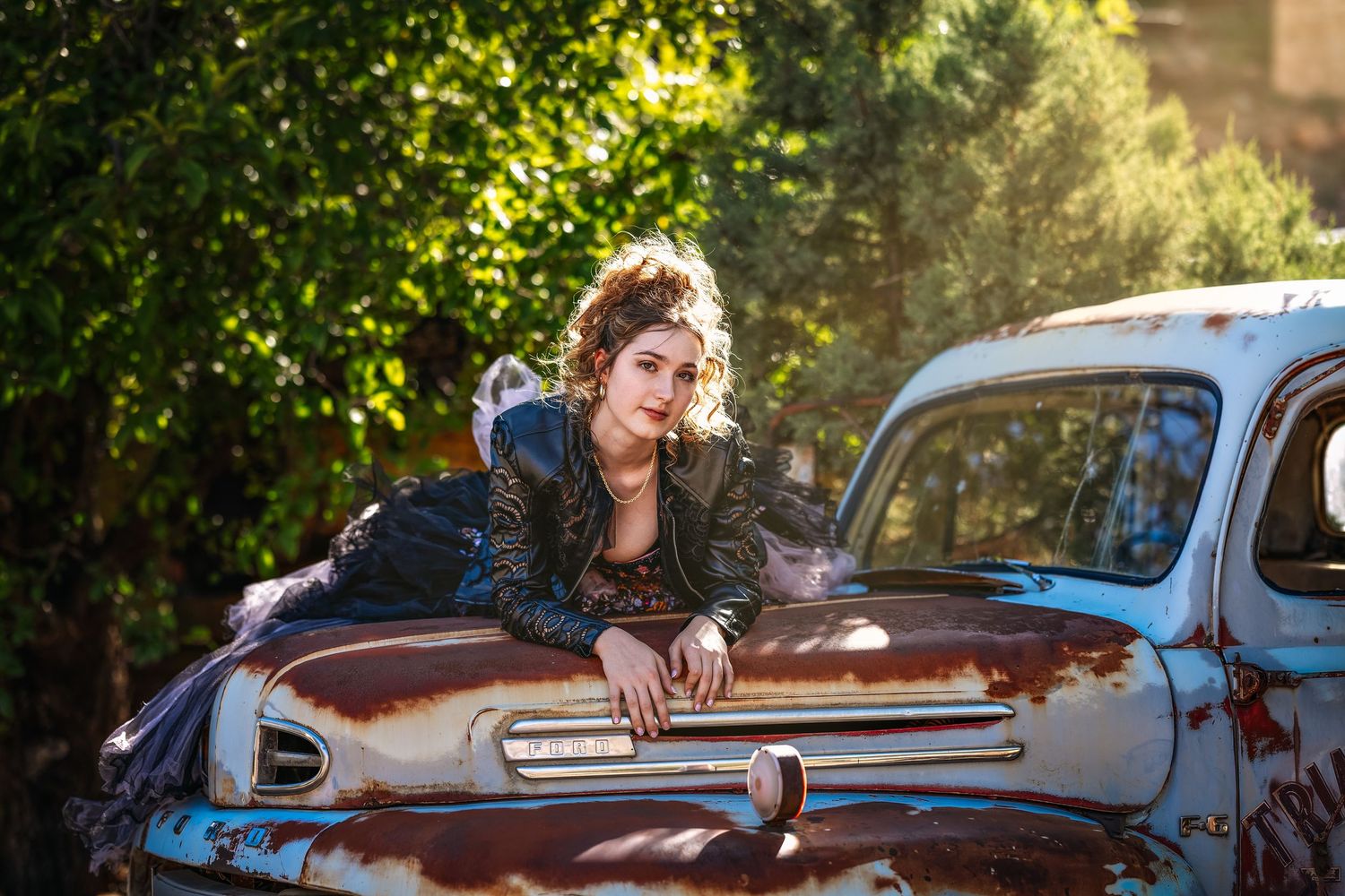 Young woman in leather jacket leaning on a rusty vintage car outdoors with sunlit green trees in background.