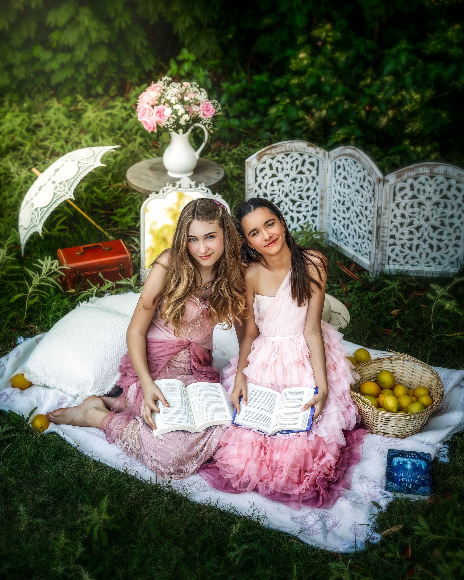 Two girls in pink tulle dresses sit on a blanket outdoors reading books with lemons and flowers nearby.