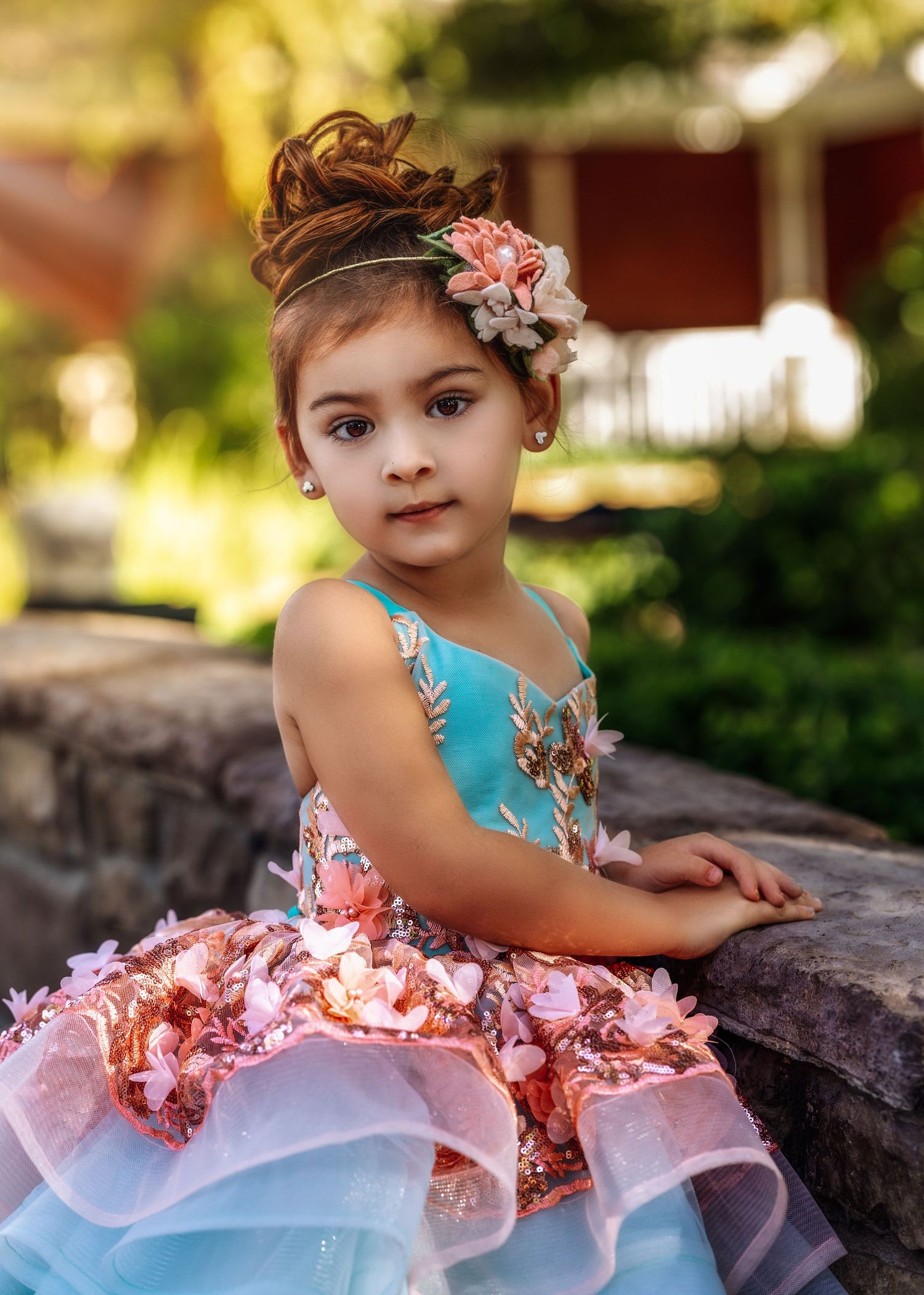 Young girl in teal and pink ruffled dress with floral headband sitting on stone wall outdoors.