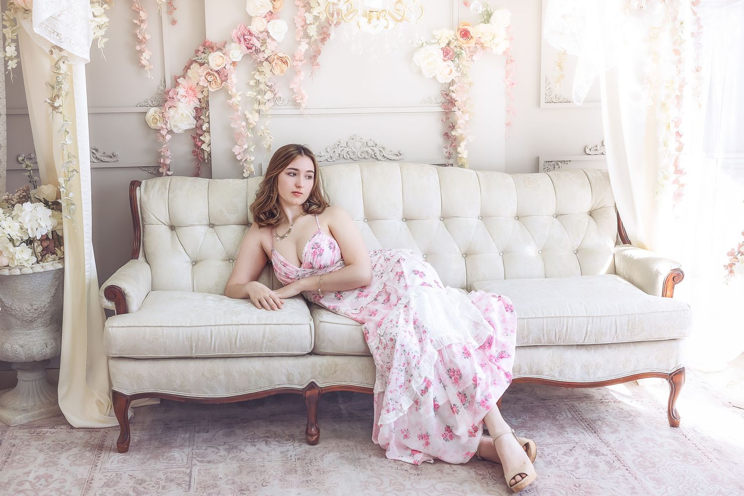 A woman in a pink floral dress reclines gracefully on a vintage white tufted sofa with floral decor.