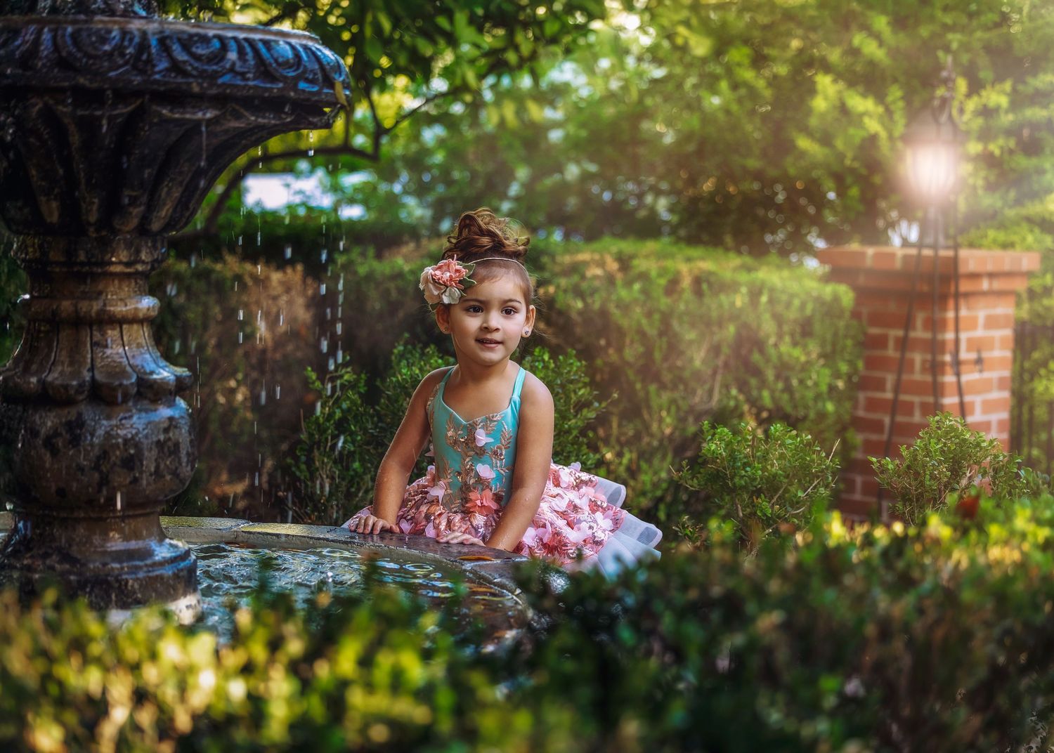 A young girl in a teal and pink tiered dress sits beside an ornate stone garden fountain in sunlight.