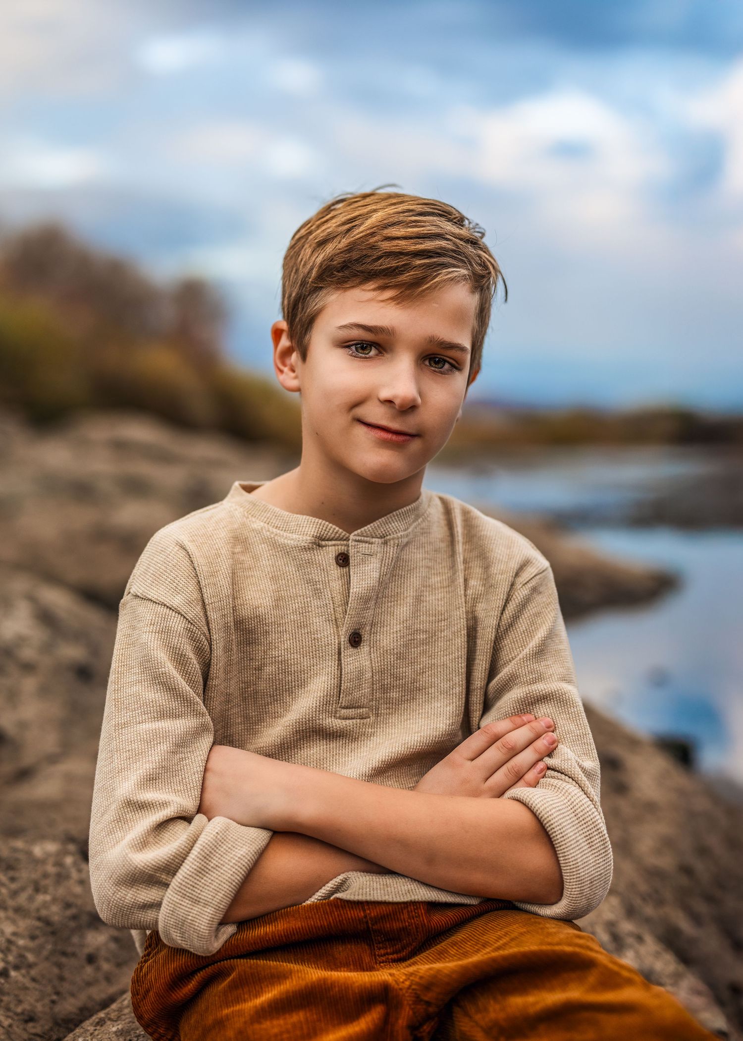 A confident young boy sits with arms crossed on rocky terrain near a river, wearing a beige henley and rust pants.