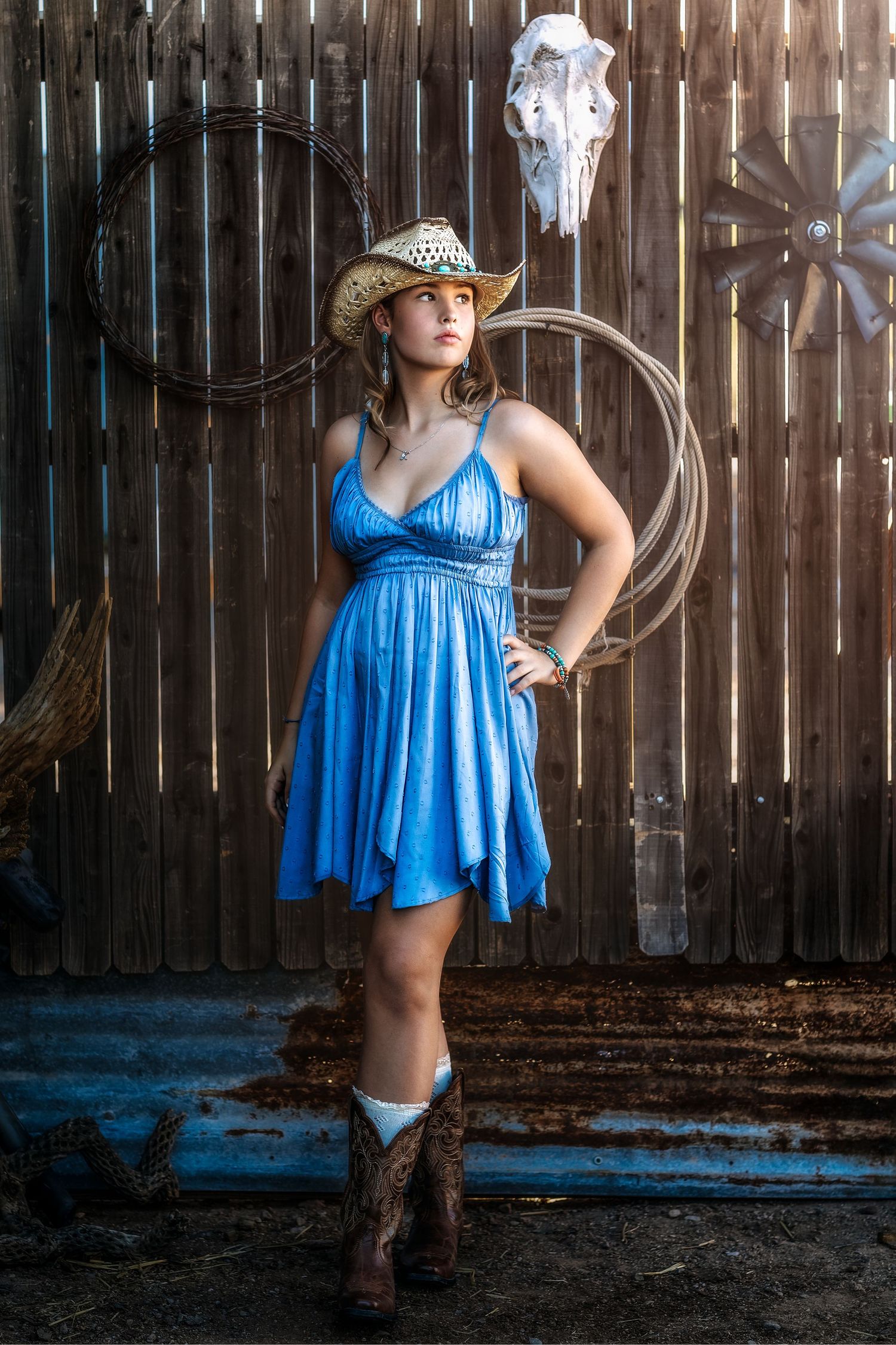 Woman in blue sundress and cowboy boots posing in rustic barn with lasso decor.