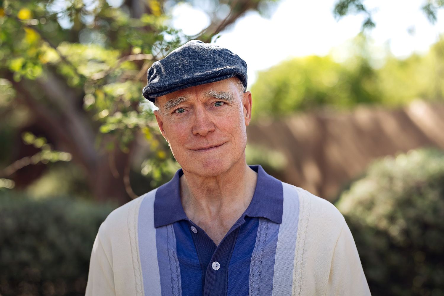 Close-up portrait of an older man in a blue striped shirt and flat cap in a sunny garden.