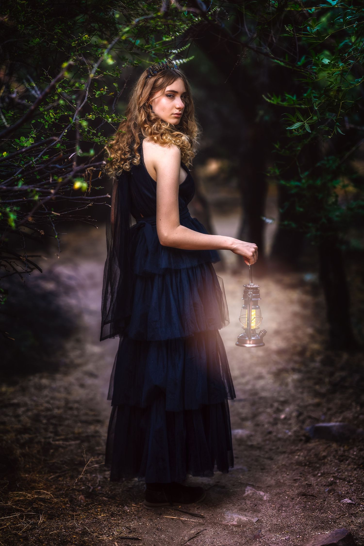 Young woman in dark blue gown holding glowing lantern on mysterious forest path at night.
