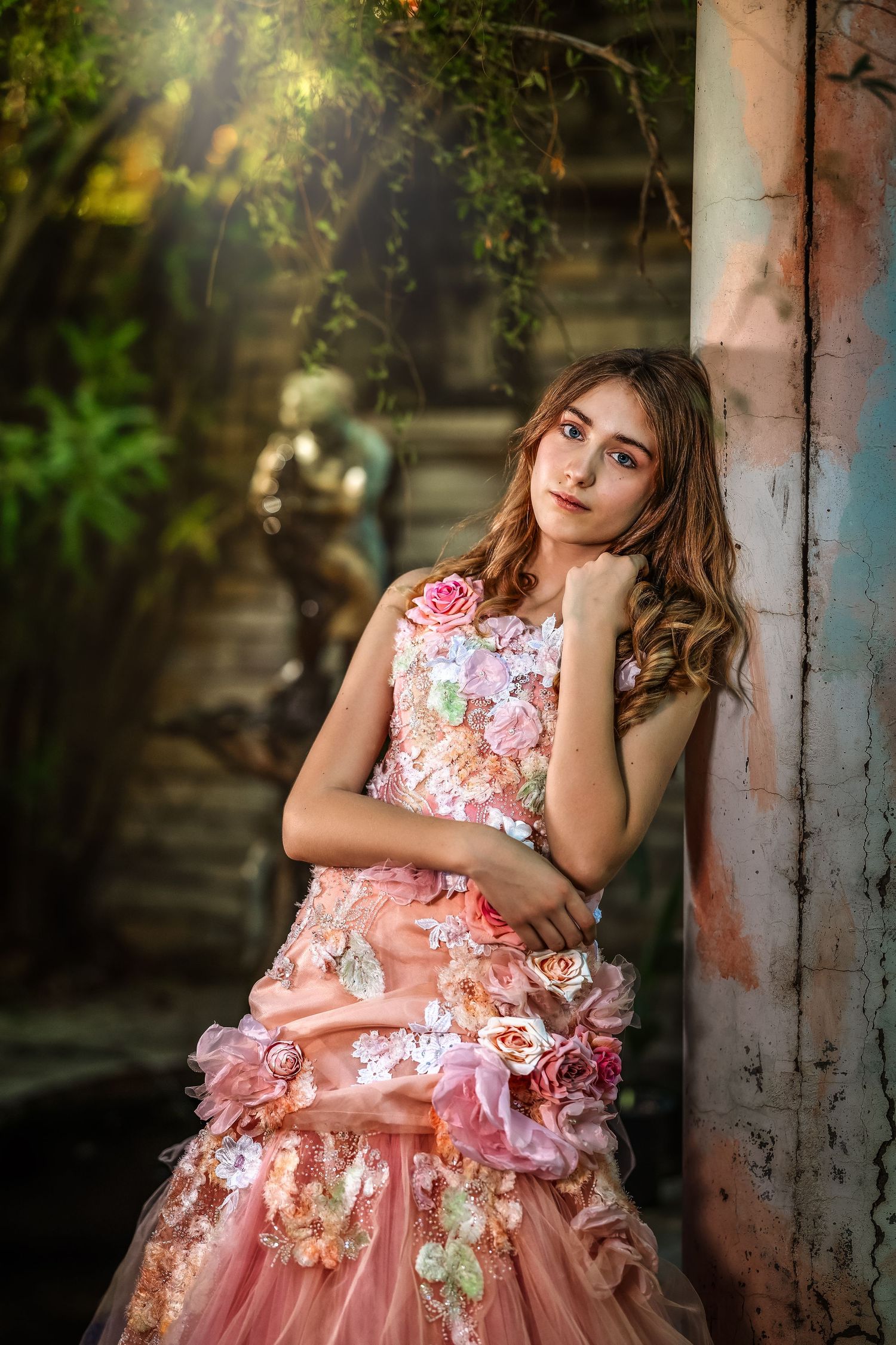 Young girl in pink floral ball gown leans against rustic column in enchanted garden setting.