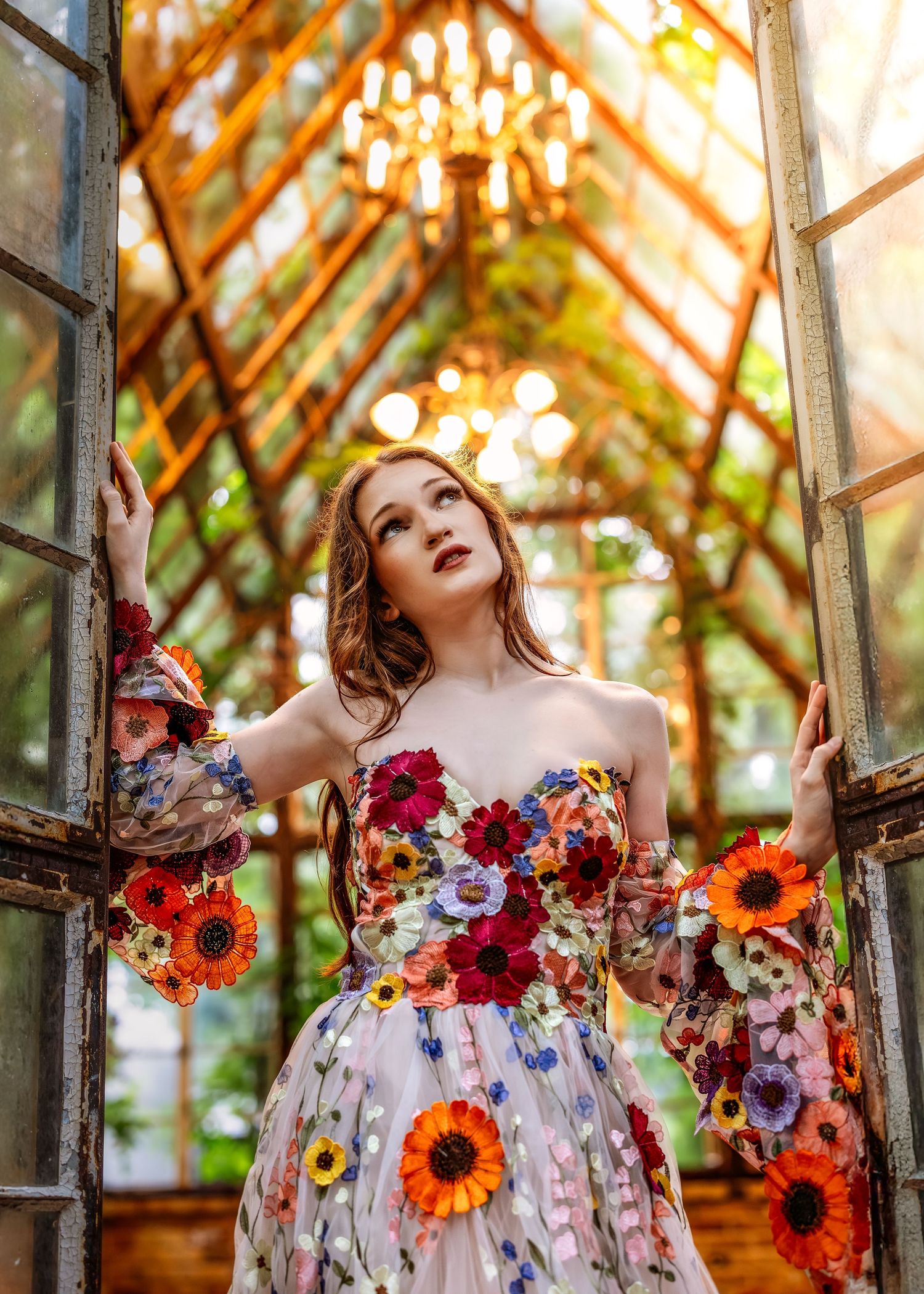 Woman in floral strapless gown poses in sunlit greenhouse doorway with chandelier overhead.