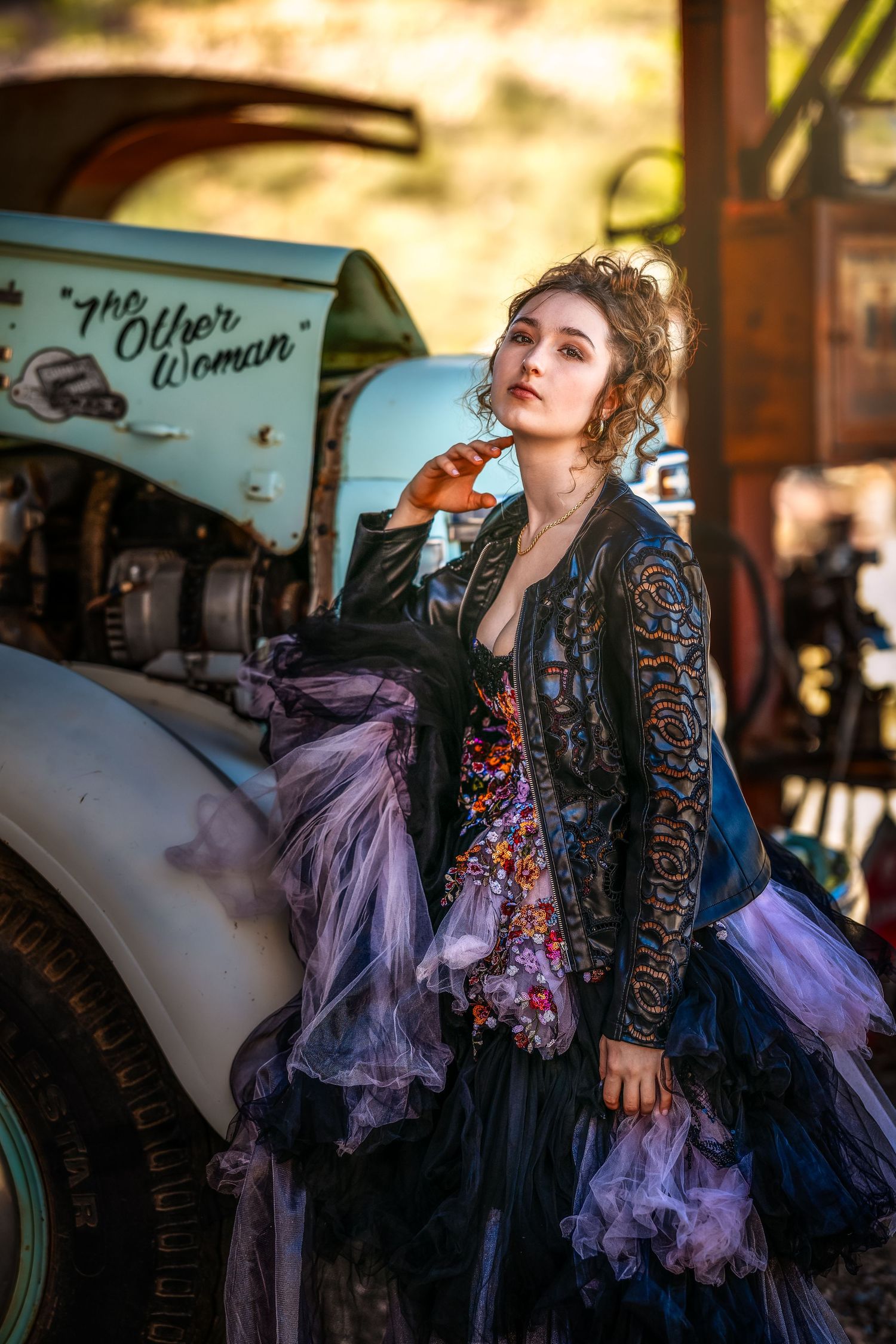 Woman in black lace jacket and purple tulle skirt poses by vintage car labeled The Other Woman outdoors.