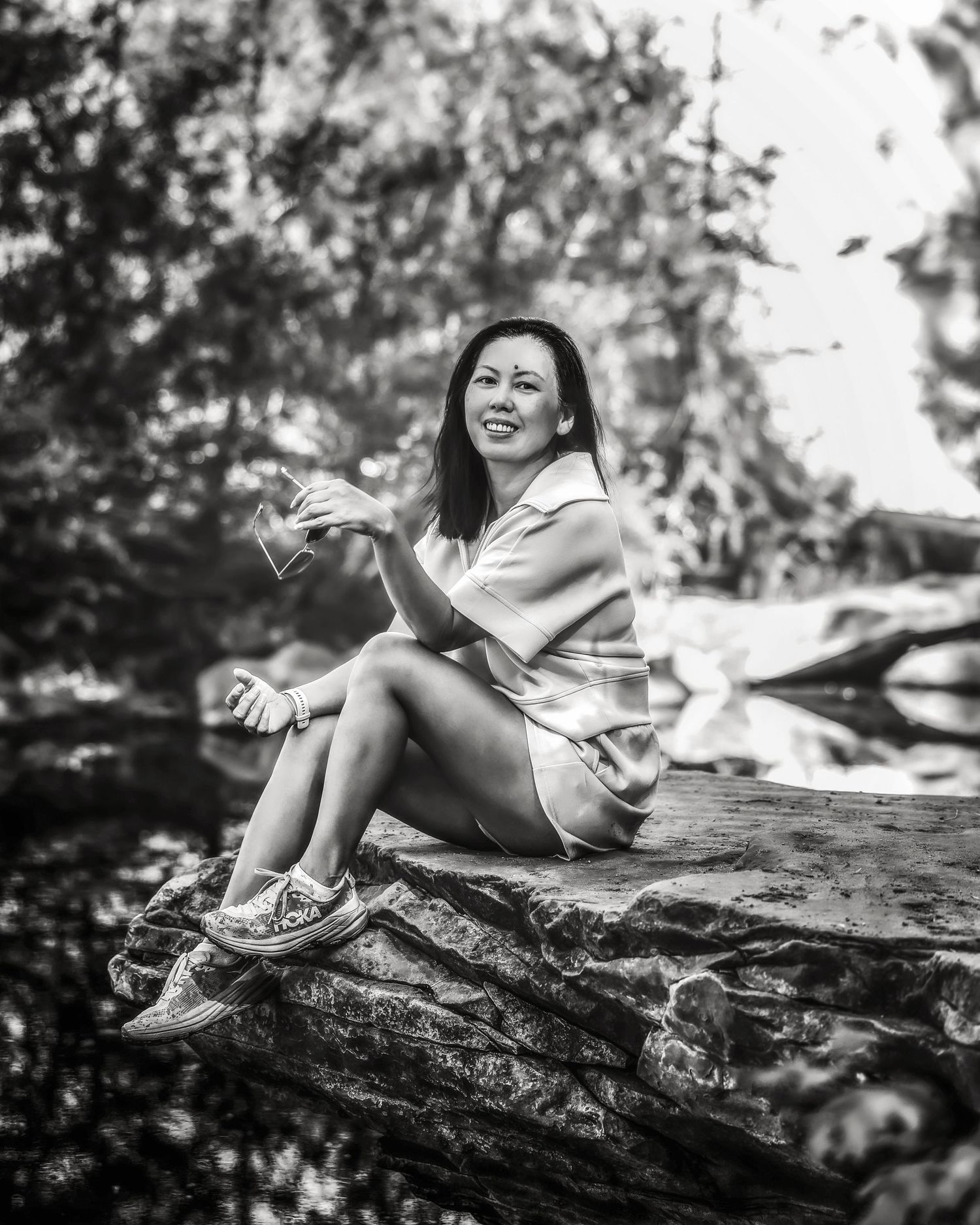 Black and white portrait of a woman sitting on a rock in a forest, holding a glass and smiling.
