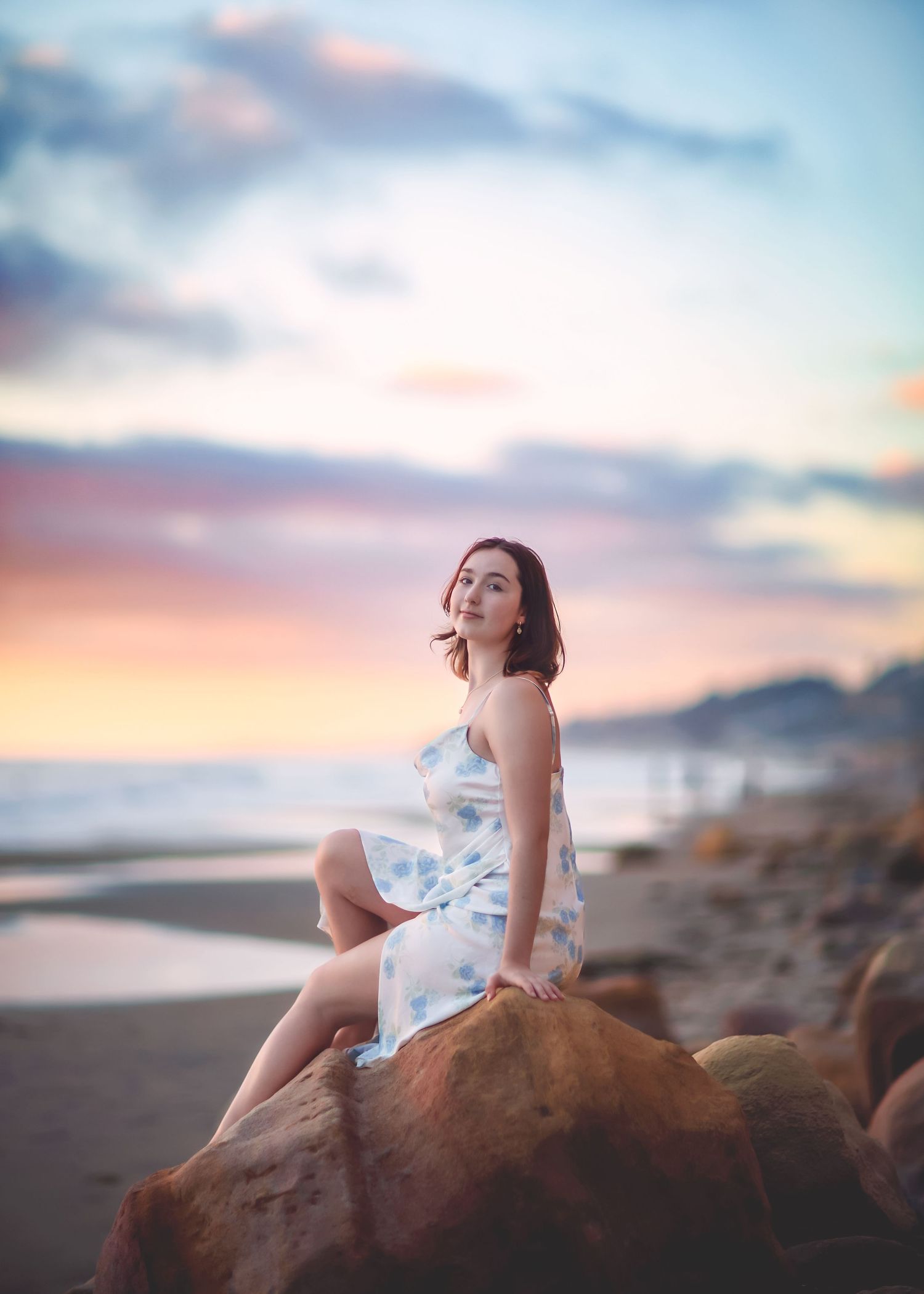 Young woman in white floral dress sitting on rock at beach sunset.