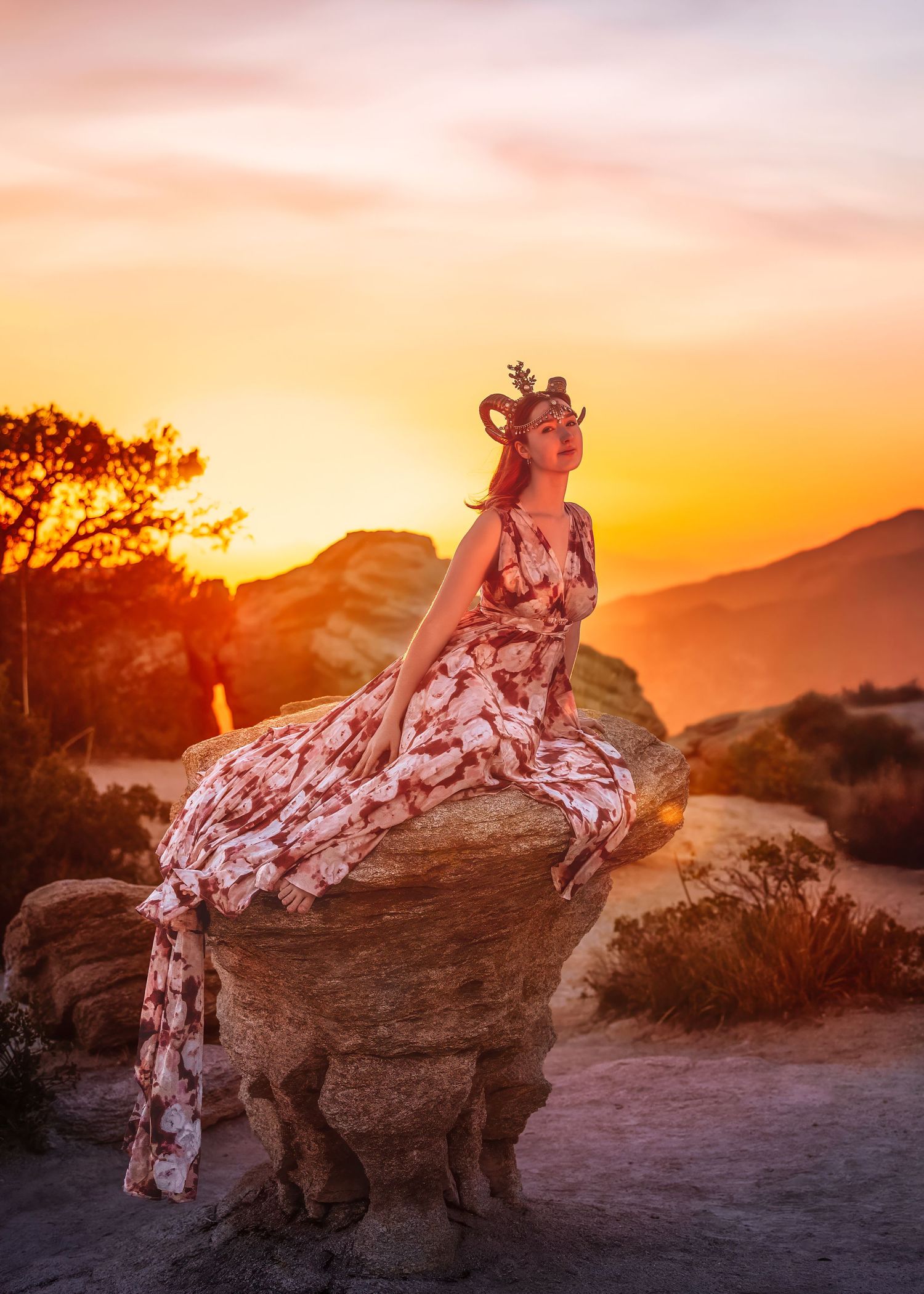 Woman in floral dress with antler crown sits on rock formation at fiery desert sunset.