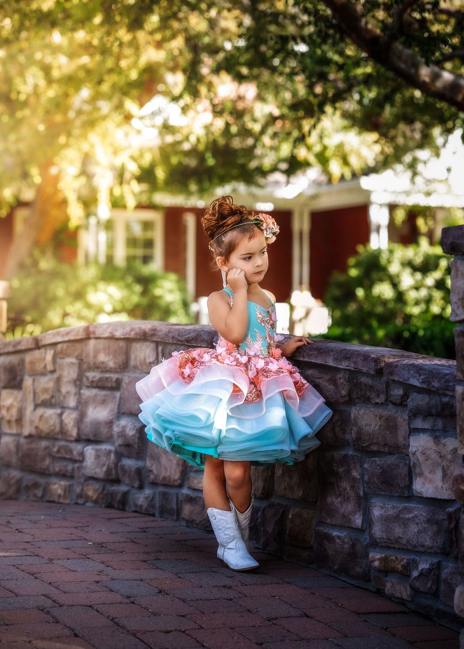 Little girl in colorful layered tutu dress with cowboy boots leaning against stone wall outdoors.