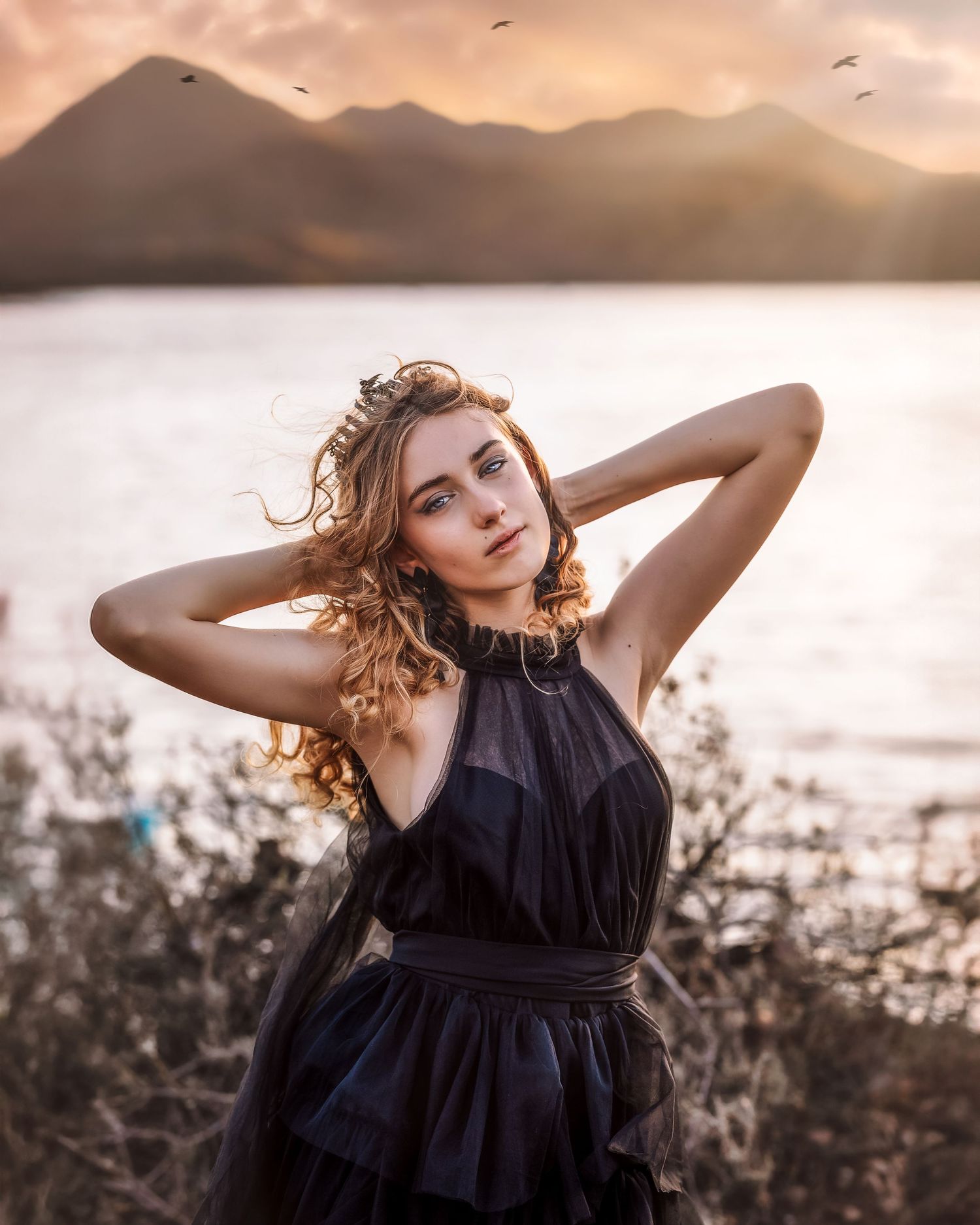 Young woman in black dress poses at golden hour with mountains and lake in the background.