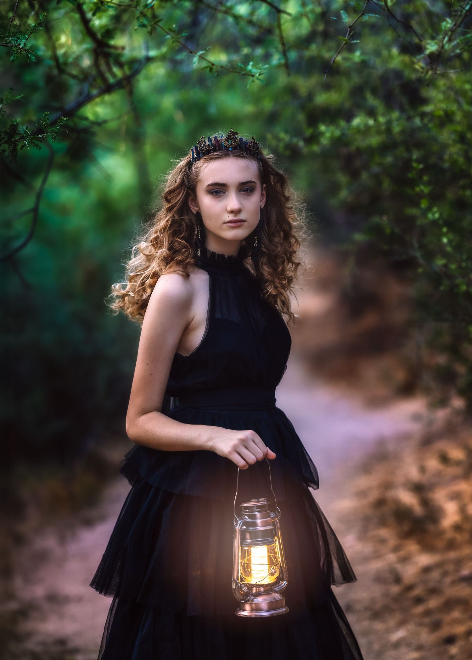 Young woman in black dress holding glowing lantern on forest path at dusk.