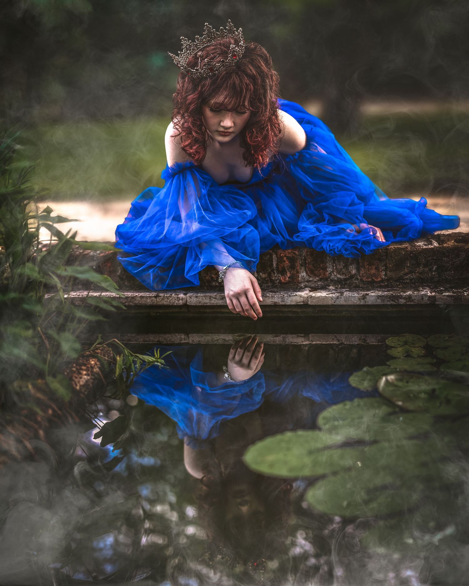 A woman in a blue gown and crown kneels by a reflective pond, her mirror image visible in the still water.