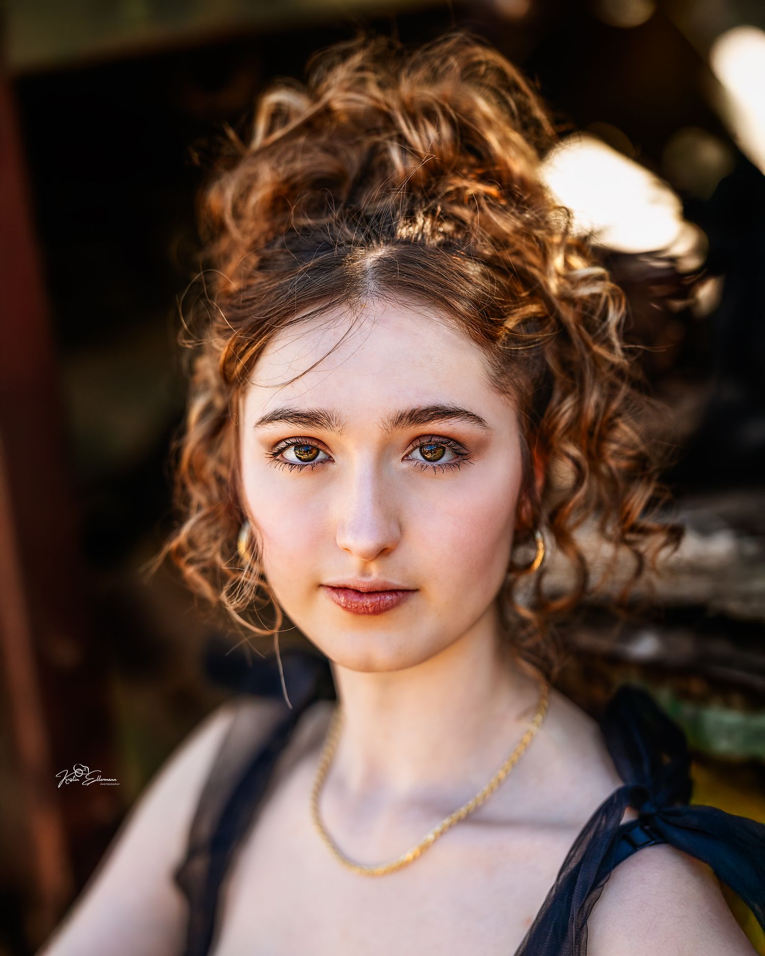 Young woman with curly auburn updo, gold hoop earrings, and necklace, posing outdoors in soft bokeh lighting.