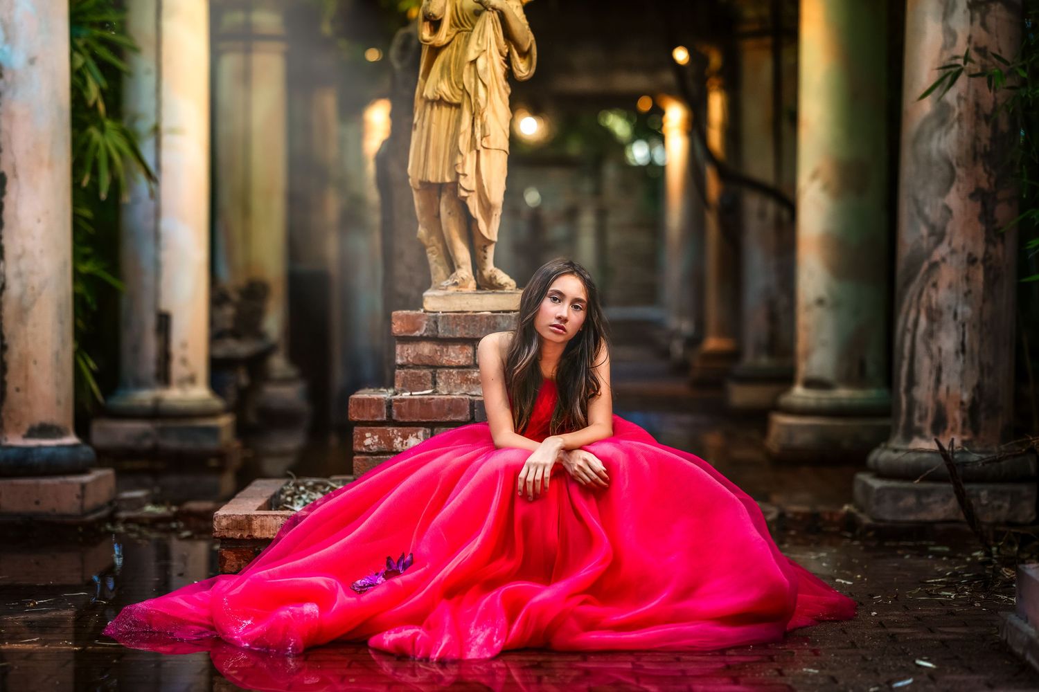 A woman in a flowing red ballgown sits gracefully on the floor near a classical statue and ornate columns.
