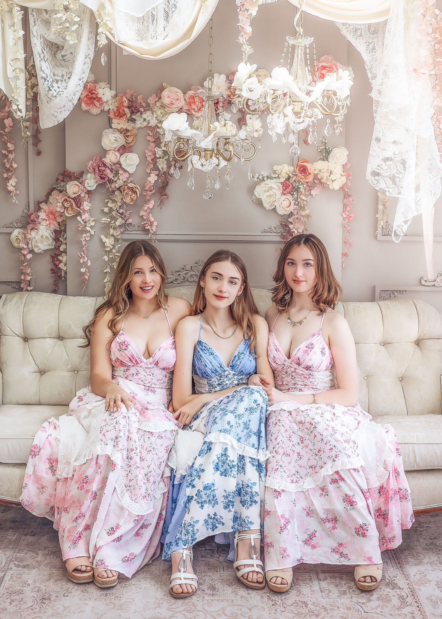 Three young women in floral maxi dresses seated on cream sofa under chandelier with flowers.