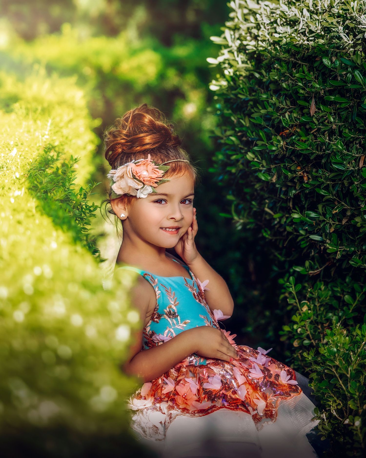 A smiling little girl in a teal floral dress with a flower headband poses between bright green garden hedges.
