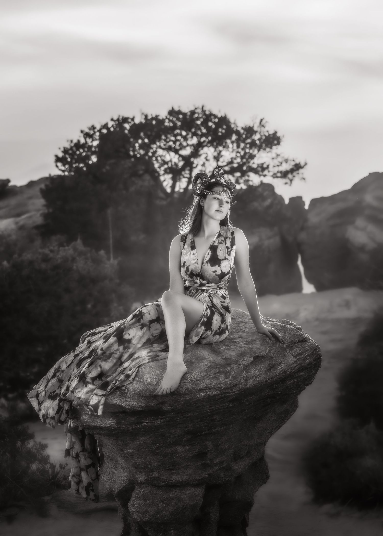 Woman in floral dress seated on mushroom rock at dusk, black and white fine art portrait.