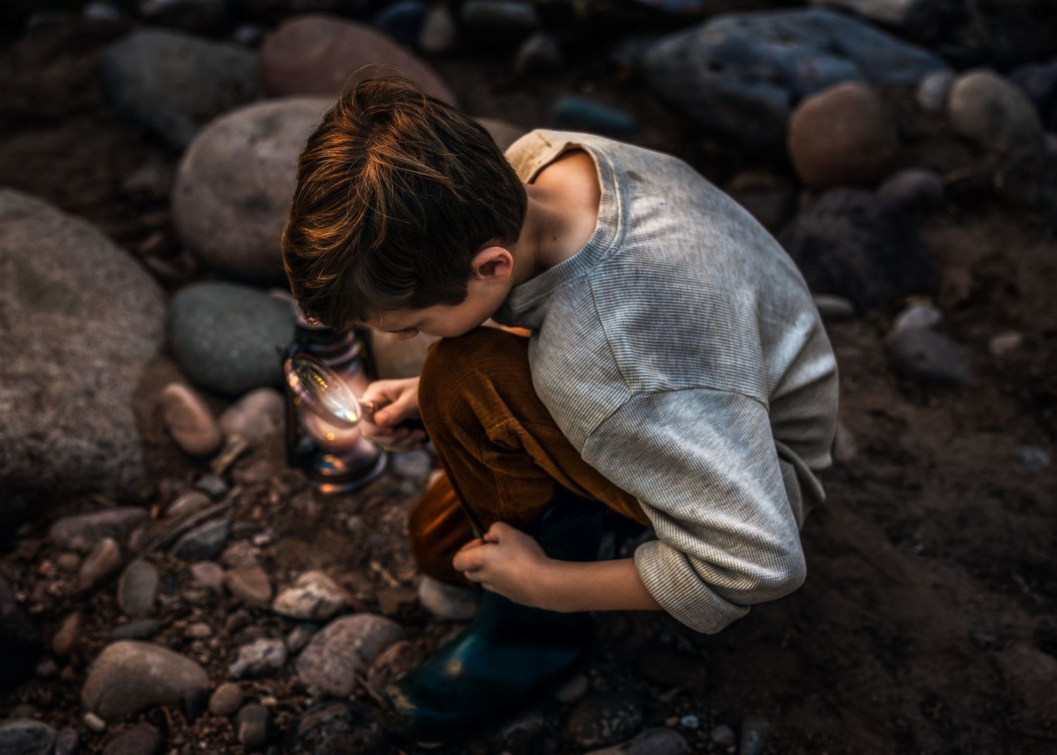 Young boy crouches among river rocks examining something closely with a magnifying glass in moody natural light.