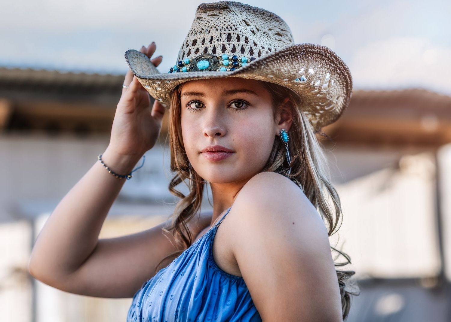 Close-up portrait of young woman in turquoise-accented cowboy hat holding brim outdoors.