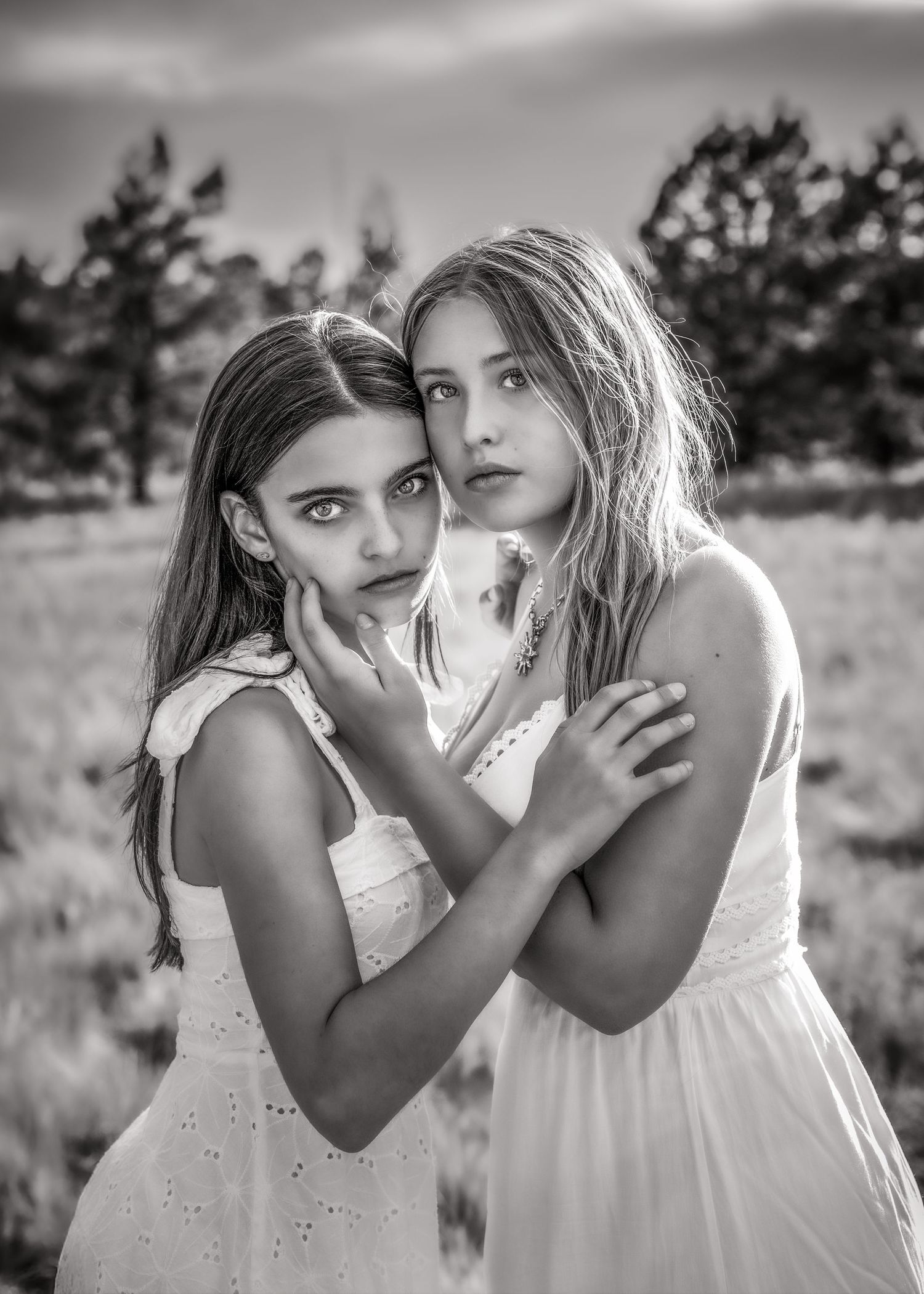 Two girls in white dresses embrace in a field at dusk in a black and white portrait photo.