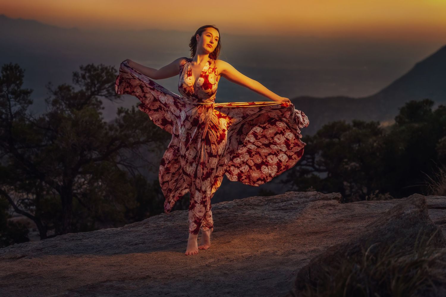 Woman in floral dress dancing on rocky mountain at sunset with misty valley backdrop.
