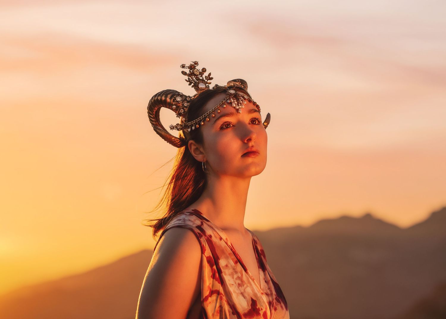 Close-up portrait of woman wearing ornate ram horn crown against glowing orange sunset sky.