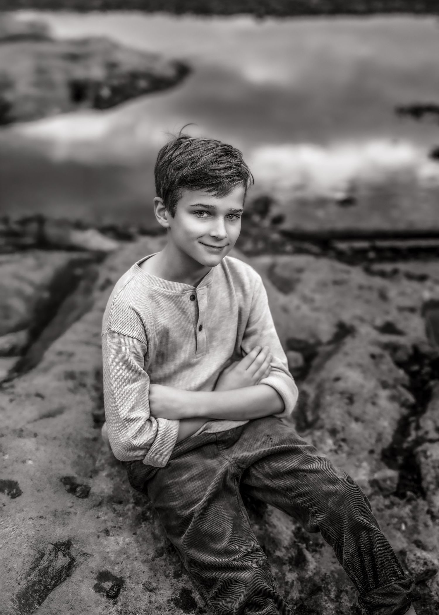 Black and white portrait of a boy sitting on rocks by water with arms crossed, smiling.