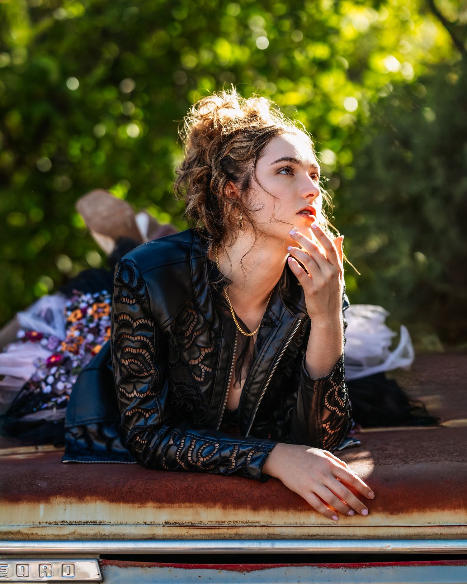 A young woman in a black lace leather jacket poses thoughtfully on a vintage car hood surrounded by lush greenery.