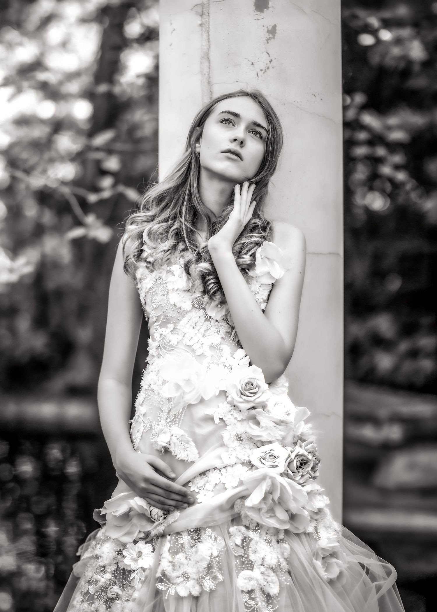 Black and white photo of a young woman in a floral wedding gown leaning against a marble column outdoors.