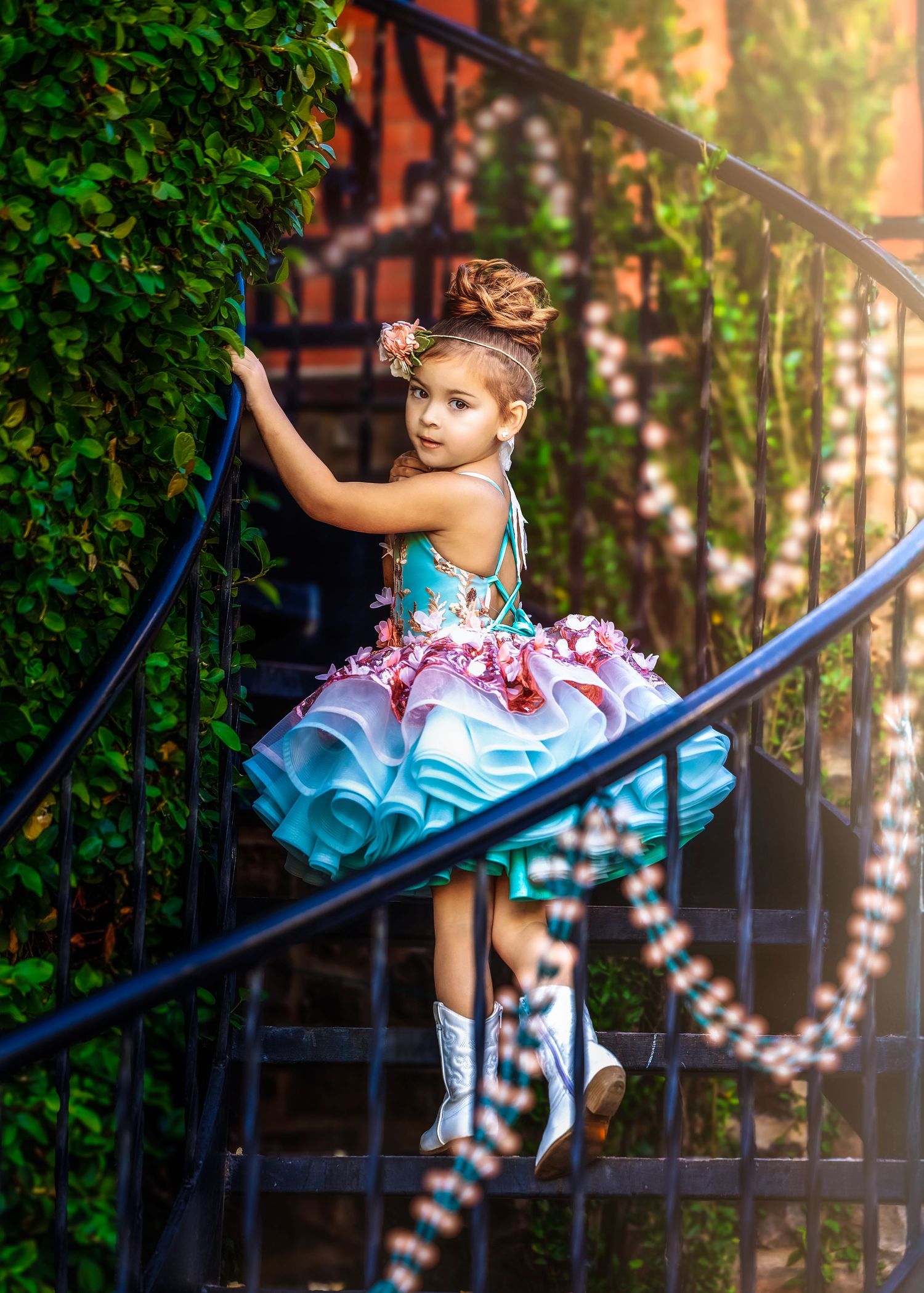 A young girl in a layered teal tutu dress and white boots poses on a spiral iron staircase with ivy backdrop.
