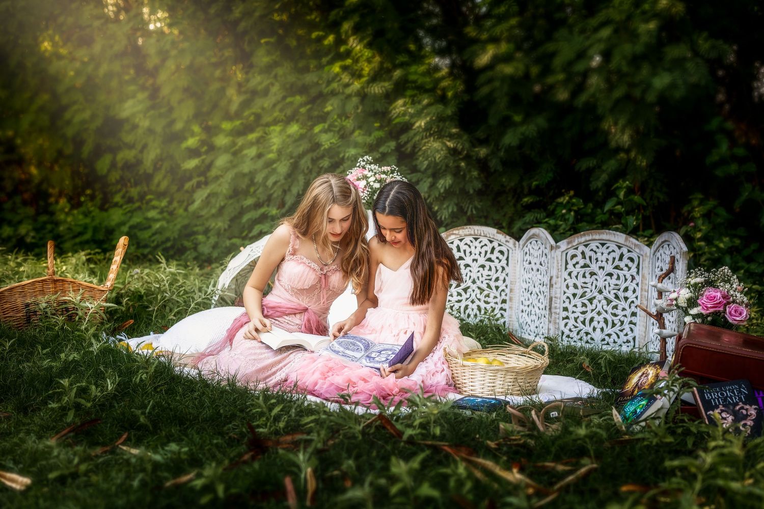 Two girls in pink dresses reading a book together on a blanket during an outdoor picnic in a sunny garden.