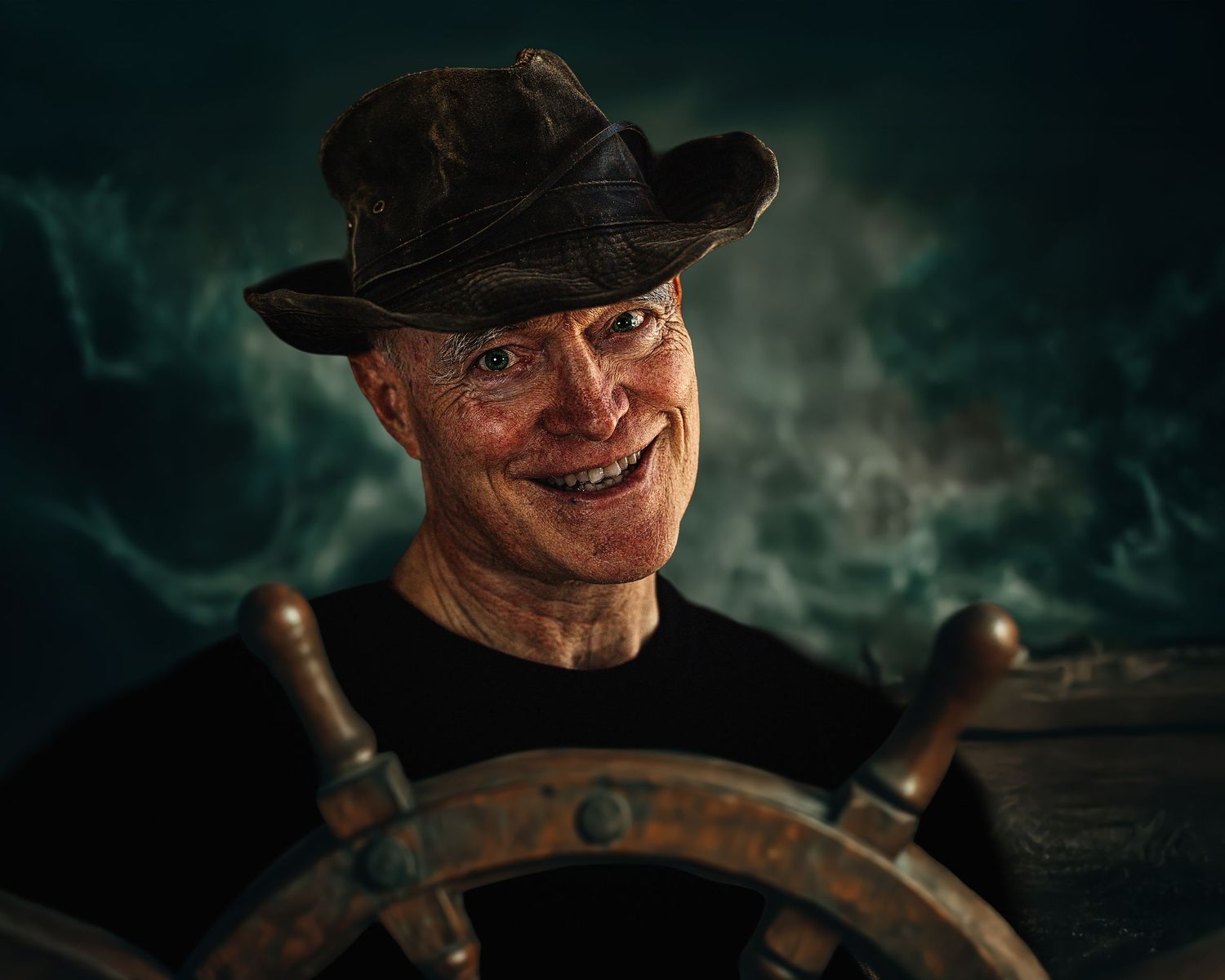 Grinning man in rugged hat standing at a ship wheel with stormy seas background.