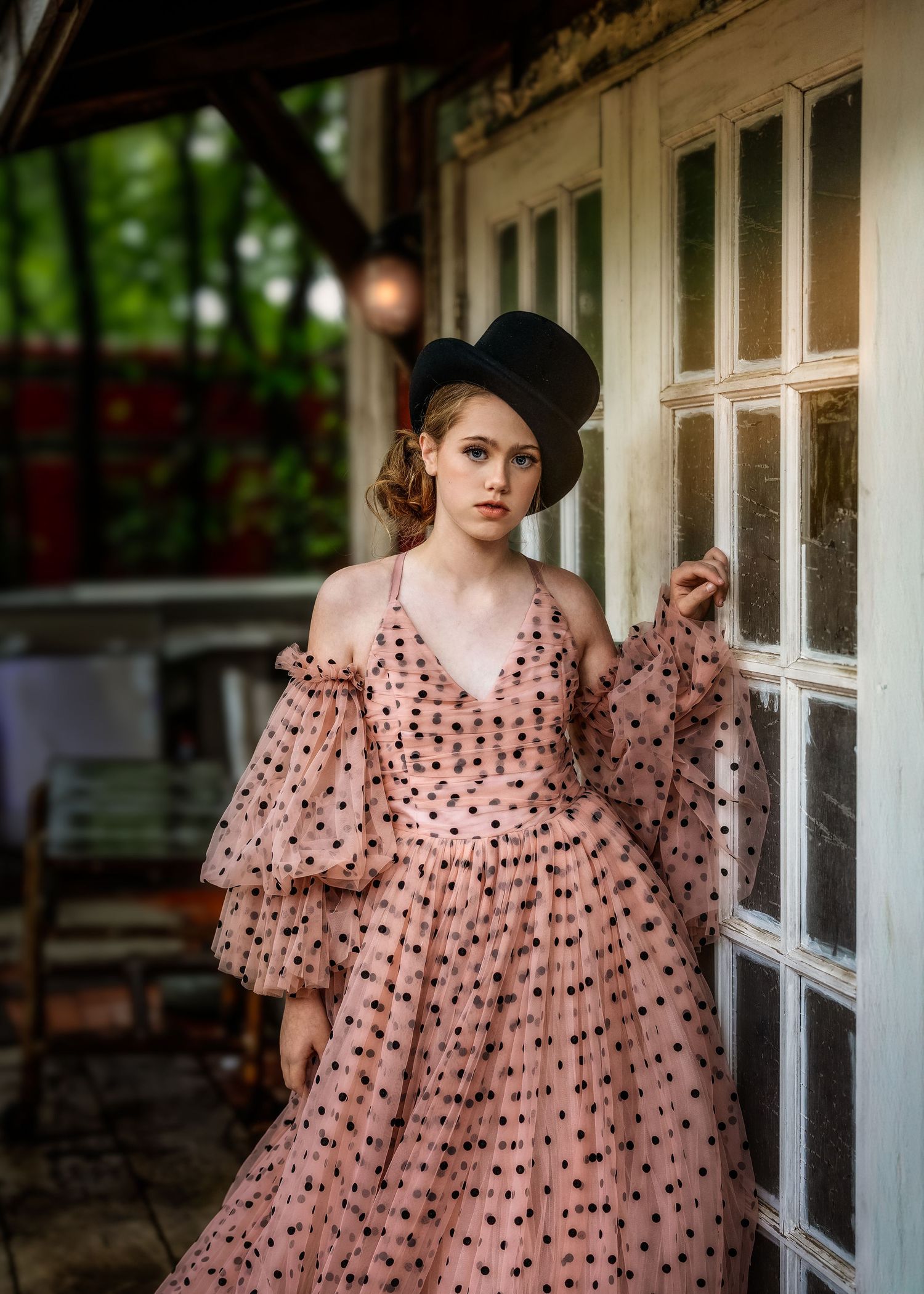 Woman in pink polka dot off-shoulder gown and black top hat poses elegantly by a vintage white door.
