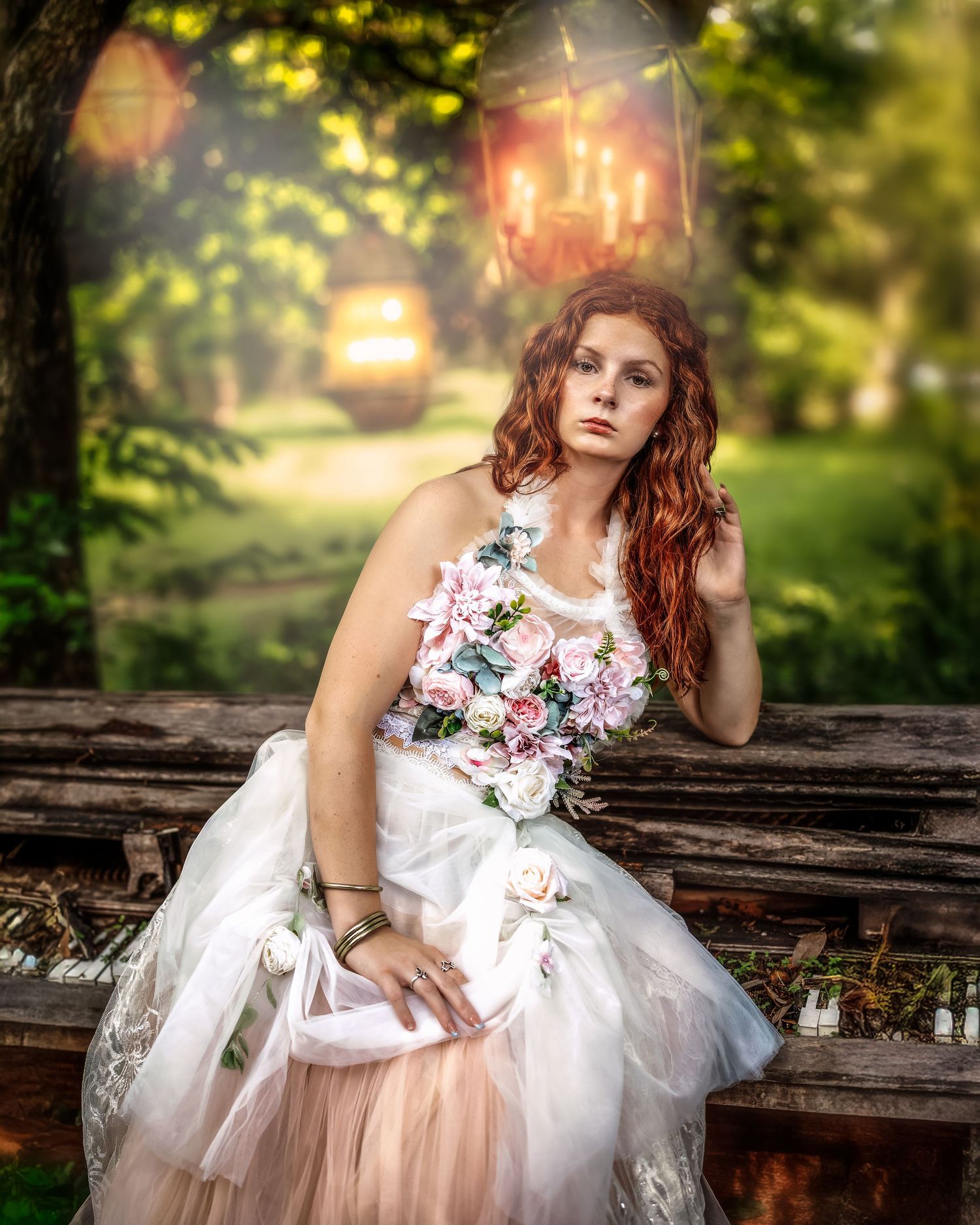 Redhead bride in floral gown holding bouquet seated outdoors with bokeh lantern lights in lush green garden.