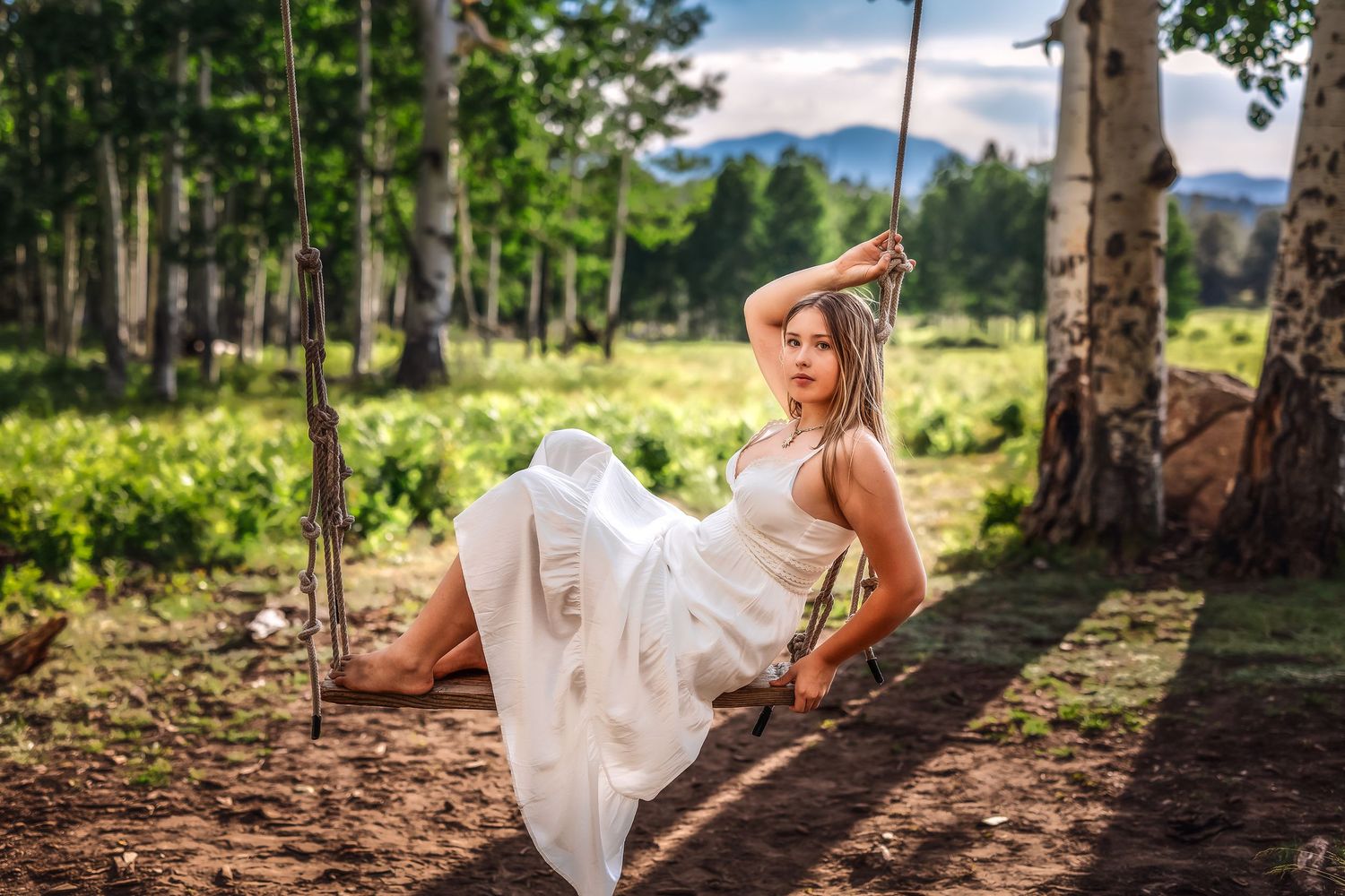 Woman in white dress relaxing on rustic rope swing in forest clearing on sunny day.