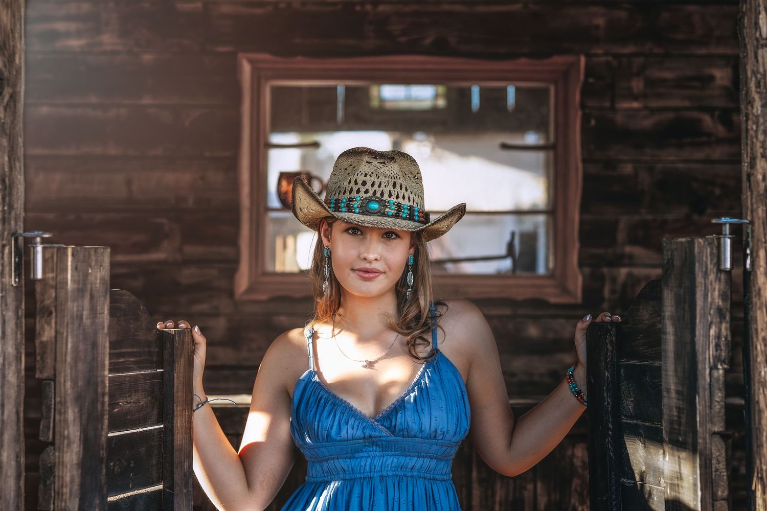 Young woman in blue dress and straw cowboy hat posing in rustic wooden doorway.