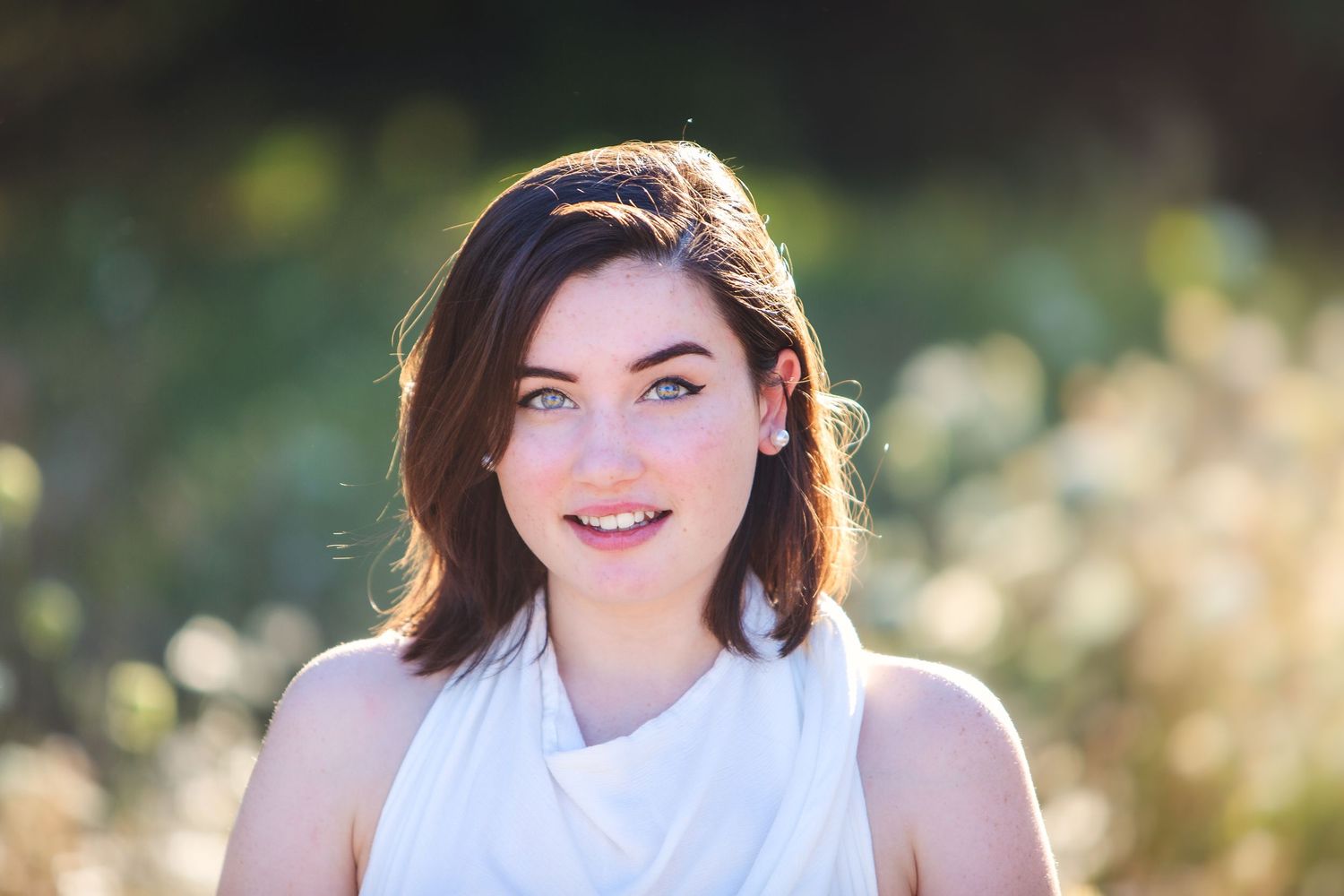 Smiling young woman with short brown hair and blue eyes in white top outdoors.