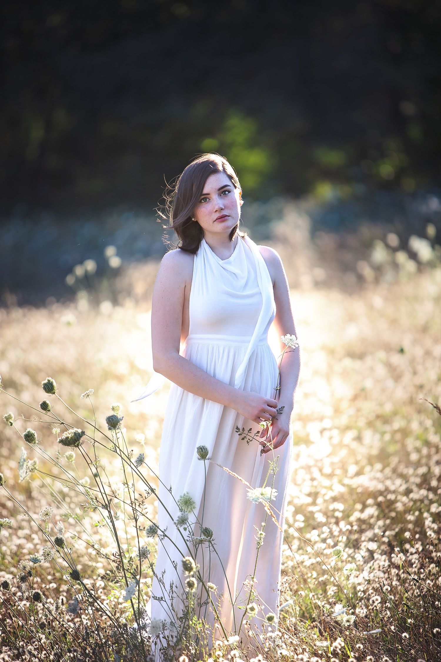 Young woman in elegant white backless dress standing among wildflowers in glowing sunlight.