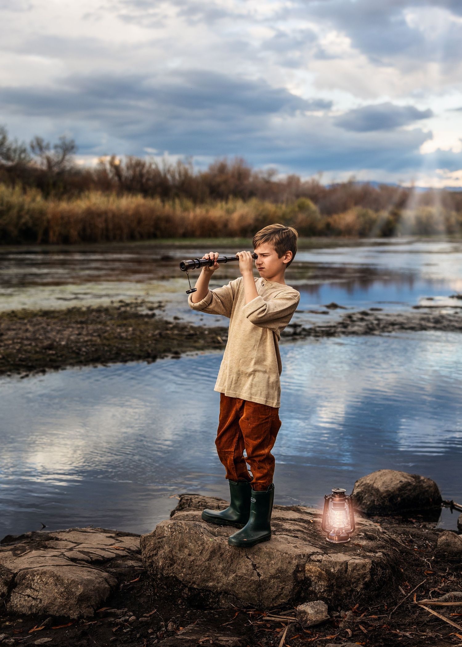 Young boy in boots stands on rocks by a lake using a telescope, with a glowing lantern beside him.