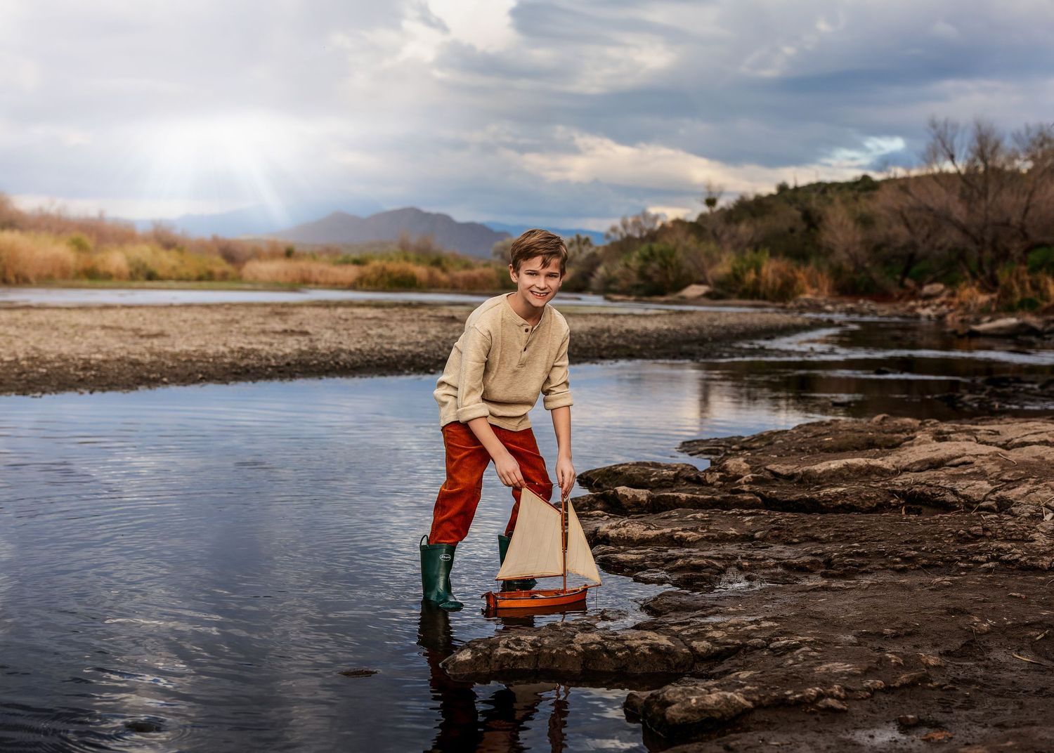 Young boy standing in a rocky river holding a wooden sailboat under dramatic cloudy skies.