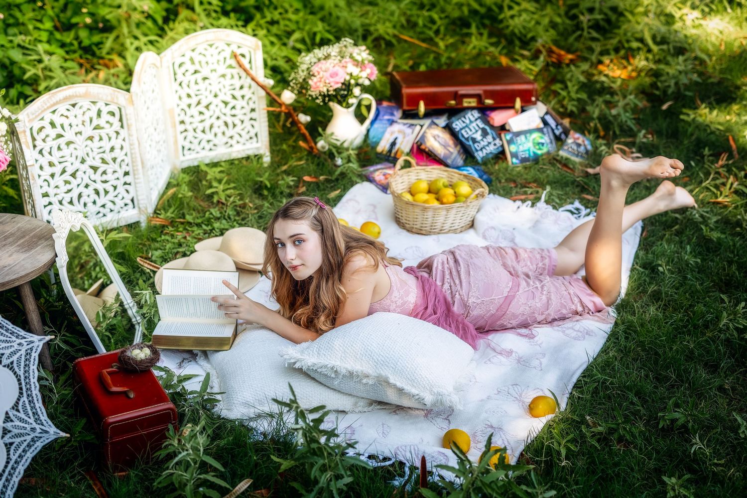 A girl in a pink dress lies on a blanket outdoors surrounded by lemons, books, and vintage garden decor.