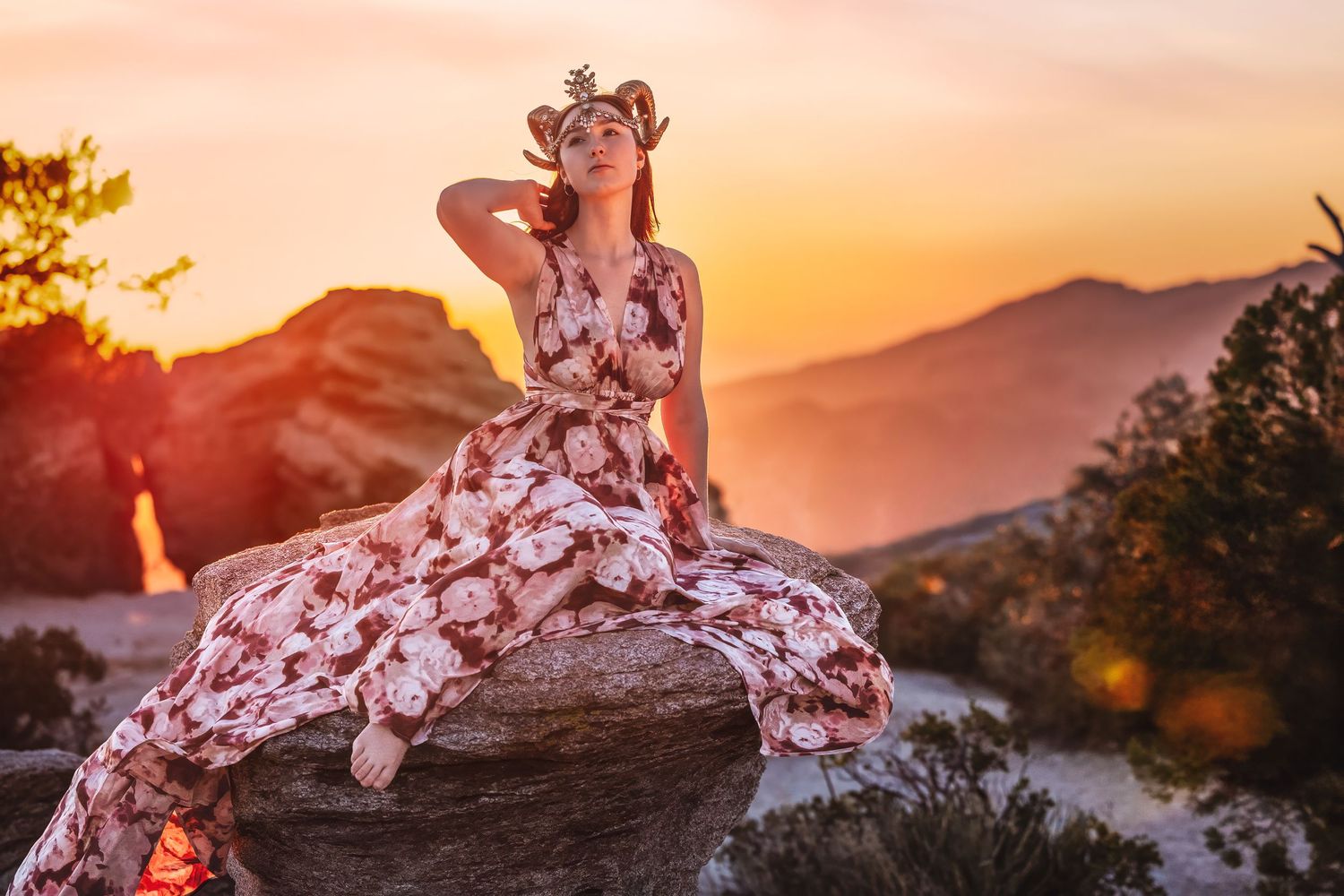 Woman in floral maxi dress with flower crown sits on rock at warm golden sunset.