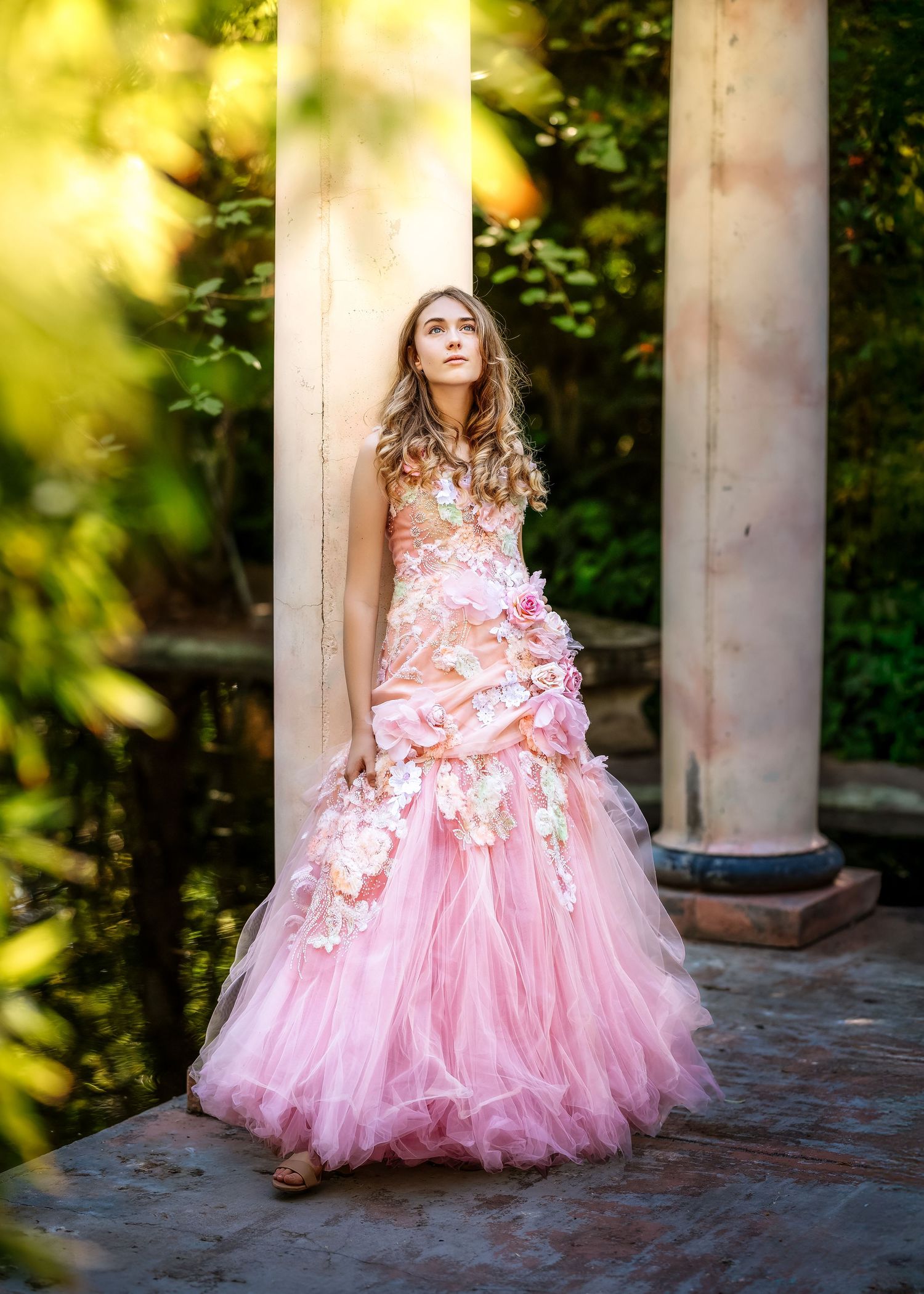 Young woman in pink floral tulle ball gown poses between white columns in a lush garden setting.