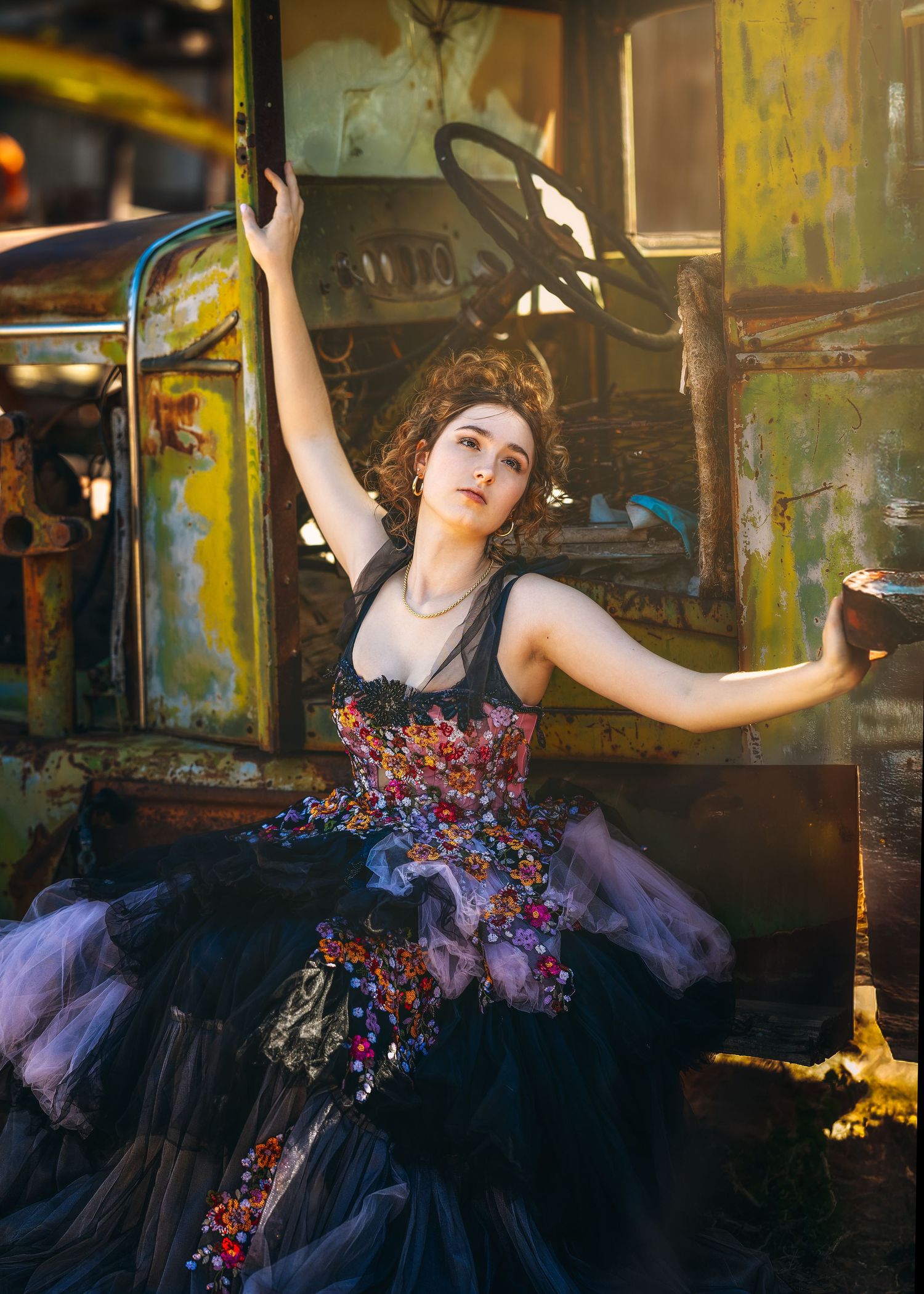 Woman in ornate black floral gown poses dramatically beside a rustic vintage vehicle in golden sunlight.