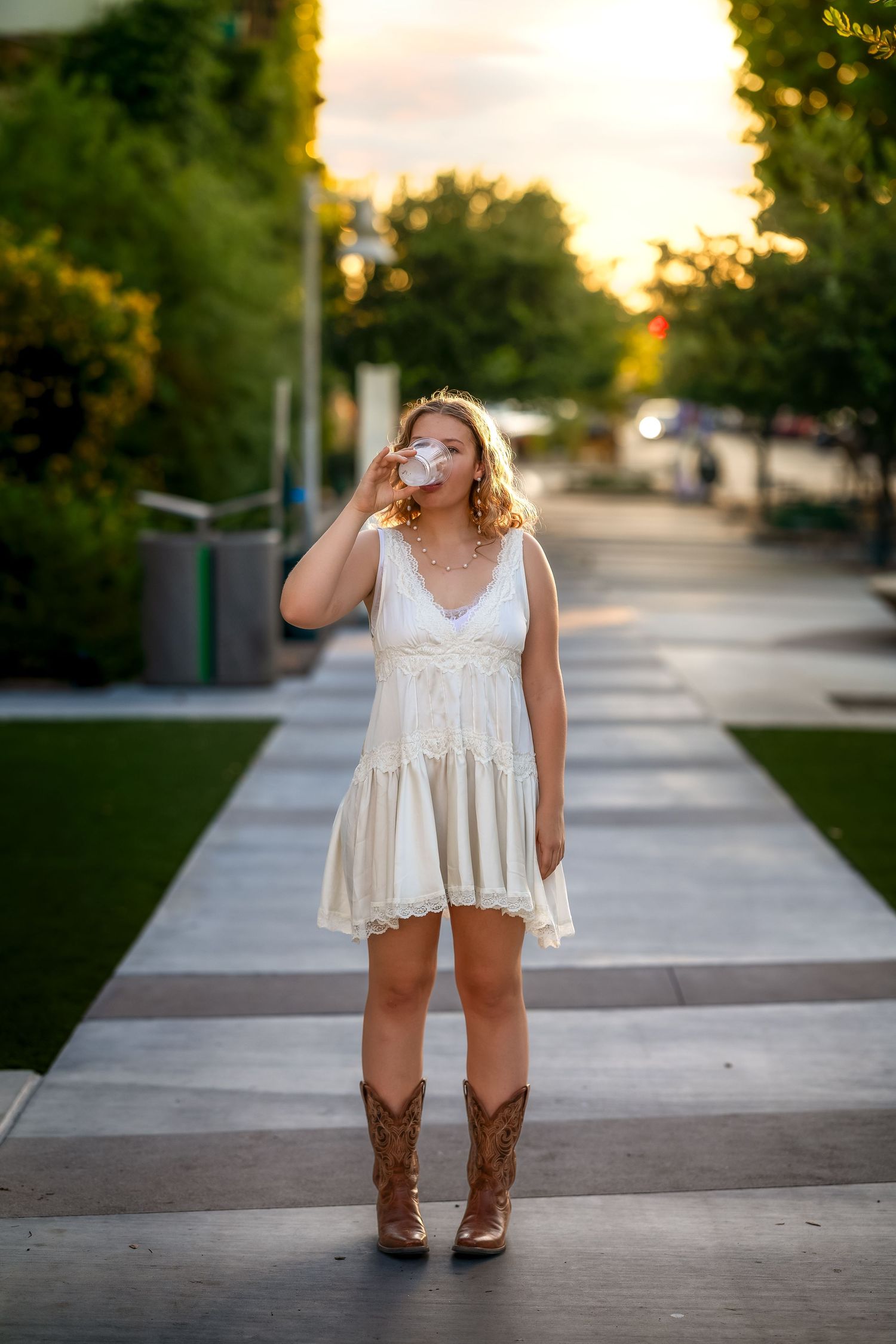Young woman in white lace dress and cowboy boots drinking from a cup on a sidewalk at sunset.